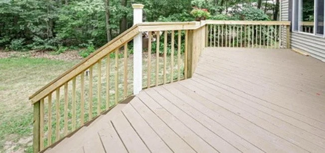 A wooden deck with railing overlooking a grassy yard and trees in the background.
