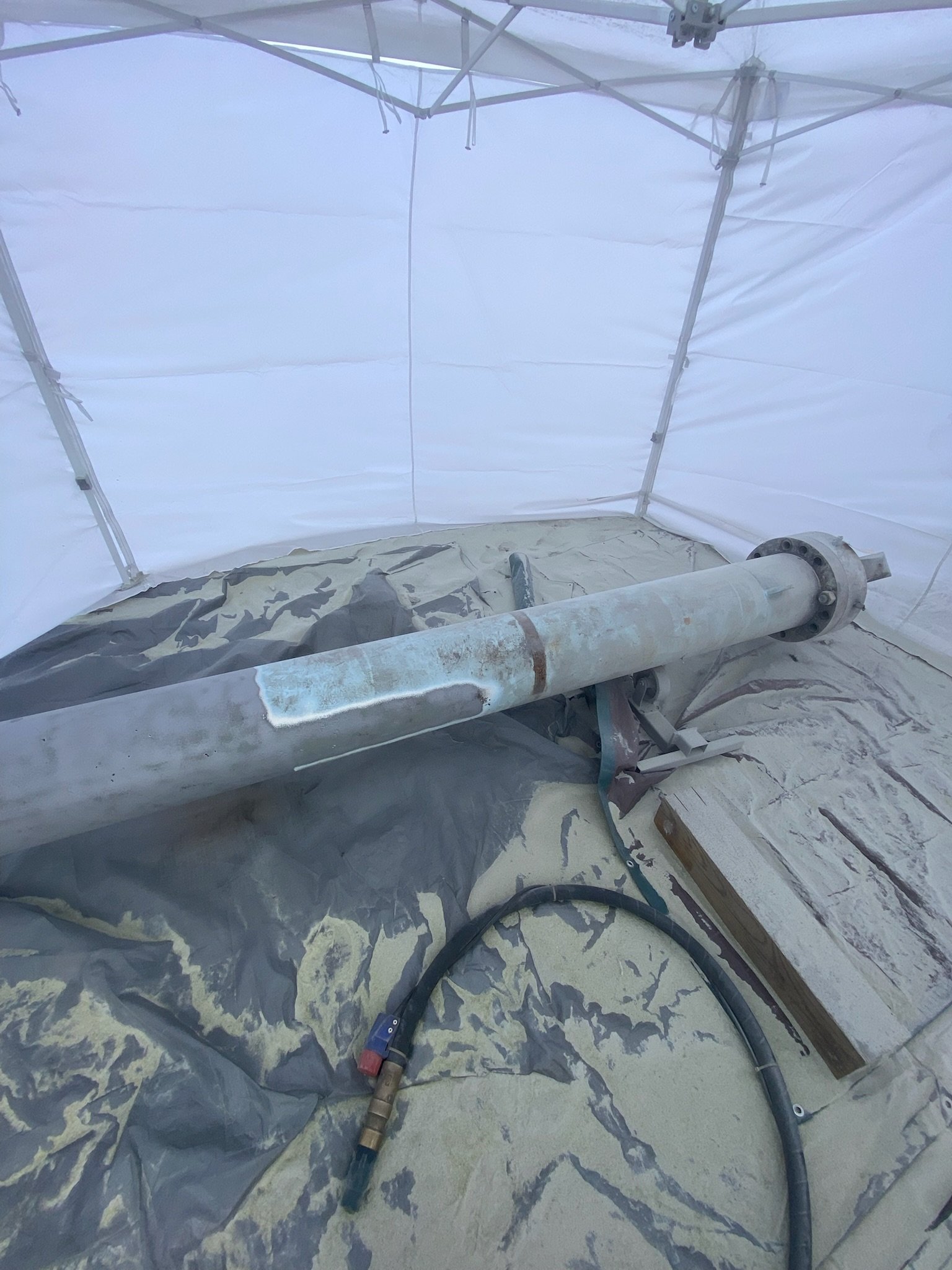 Rusty metal pipe installed on the ground inside a white containment tent with plastic sheeting and a black hose nearby.