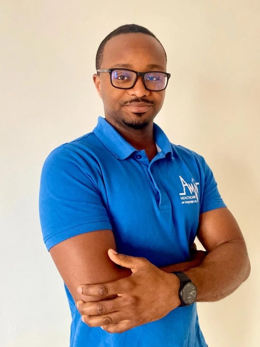 Man in een blauwe polo met een logo en de tekst 'Healthcare' op de borst, met bril, armen gekruist, staand voor een licht gekleurde muur.