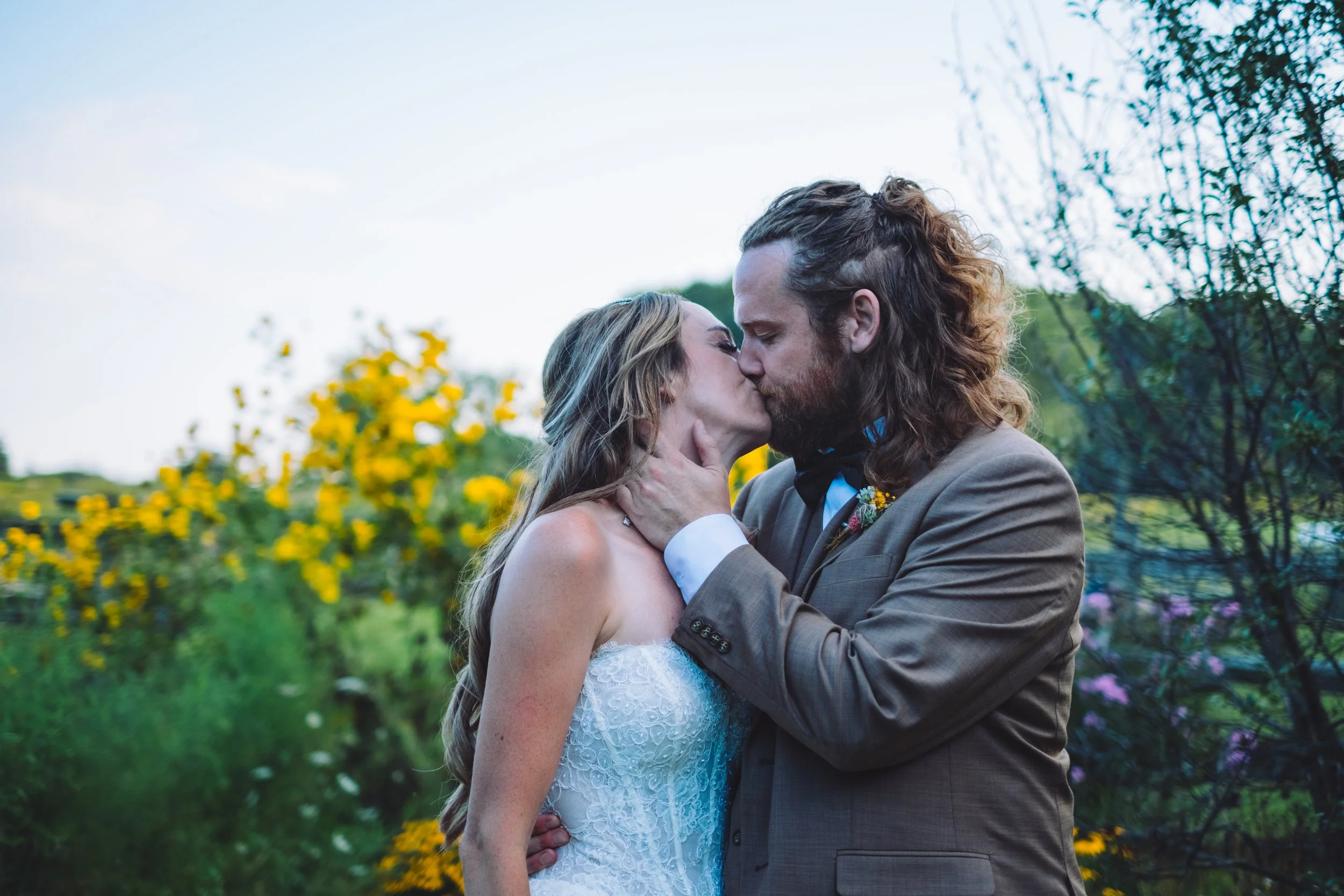 A couple sharing a kiss outdoors during a wedding, with the bride in a white lace dress and the groom in a brown suit, surrounded by yellow flowers and greenery.