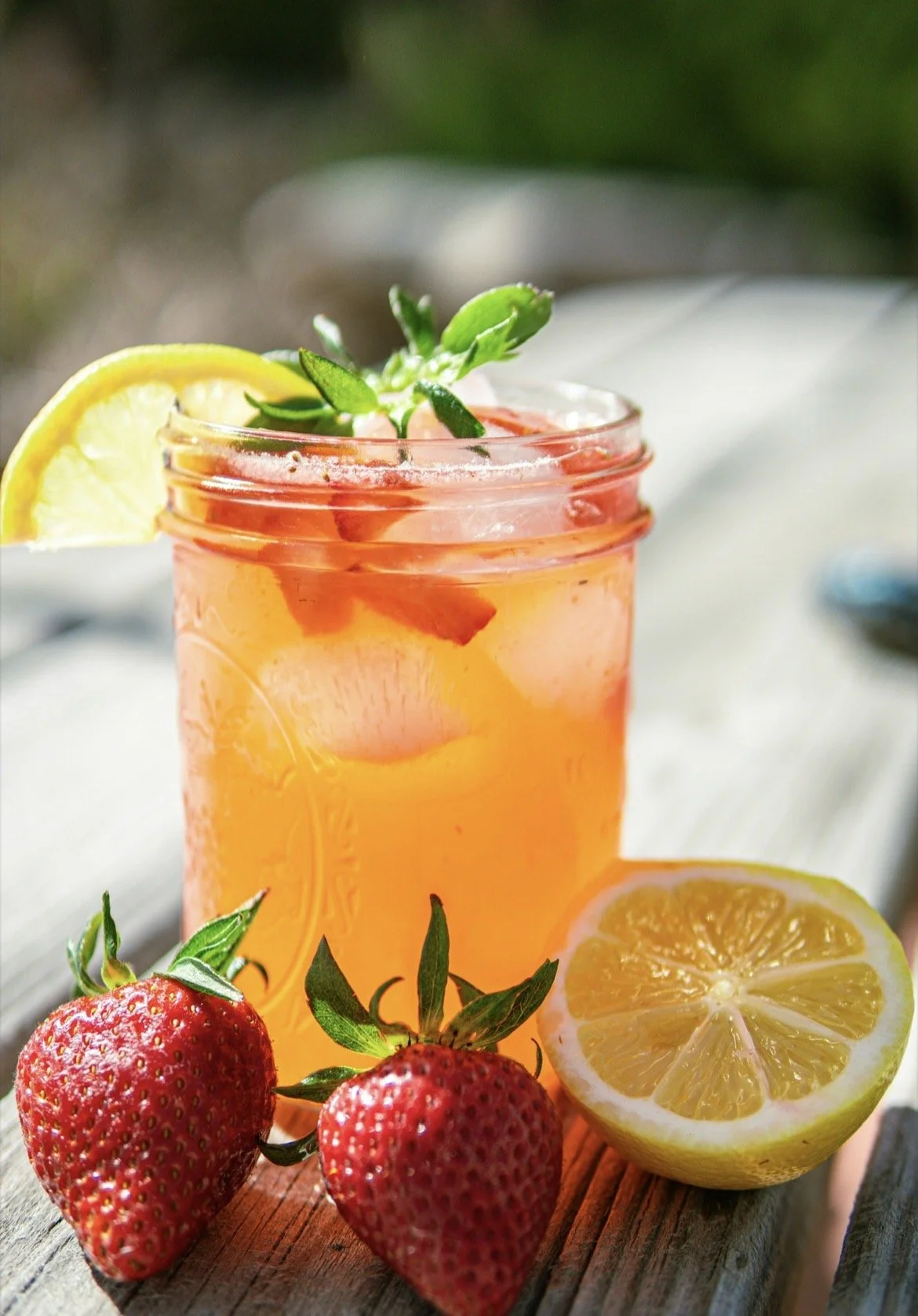 A glass jar filled with a pink-colored strawberry lemonade garnished with lemon slices, strawberry pieces, and fresh herbs. Fresh strawberries and lemon halves are placed on a rustic wooden surface beside the jar.