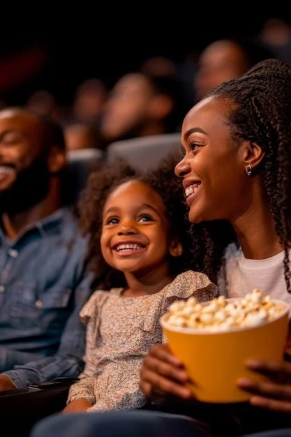 A woman and young girl sitting in a movie theater, smiling and watching a film, with the woman holding a yellow bucket of popcorn.