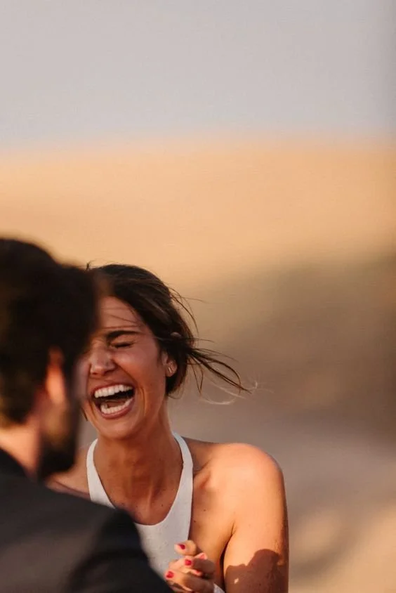 A woman with short hair, smiling and laughing, wearing a white sleeveless top, outdoors during sunset, with a blurry man in the foreground.
