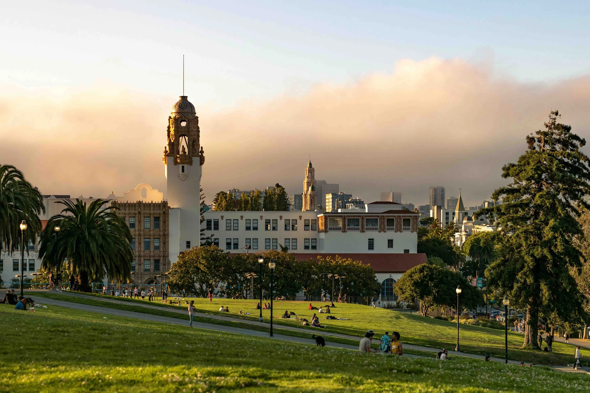 Sunset over a city park with green grass, trees, people sitting and walking, historic buildings, and city skyline in the background.