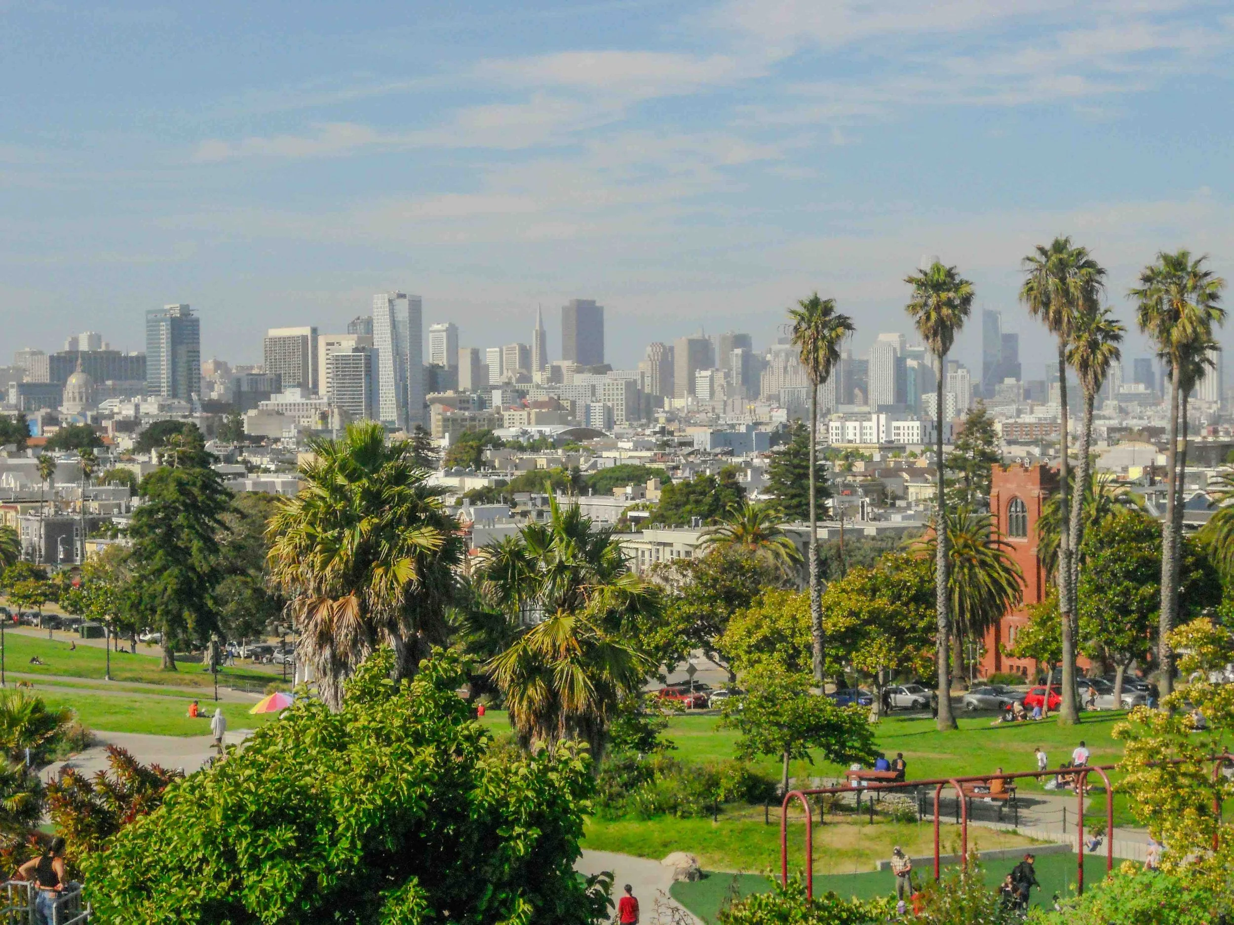 A city skyline with tall buildings and skyscrapers, foregrounded by a park with green trees and tall palm trees, and several people walking and relaxing.