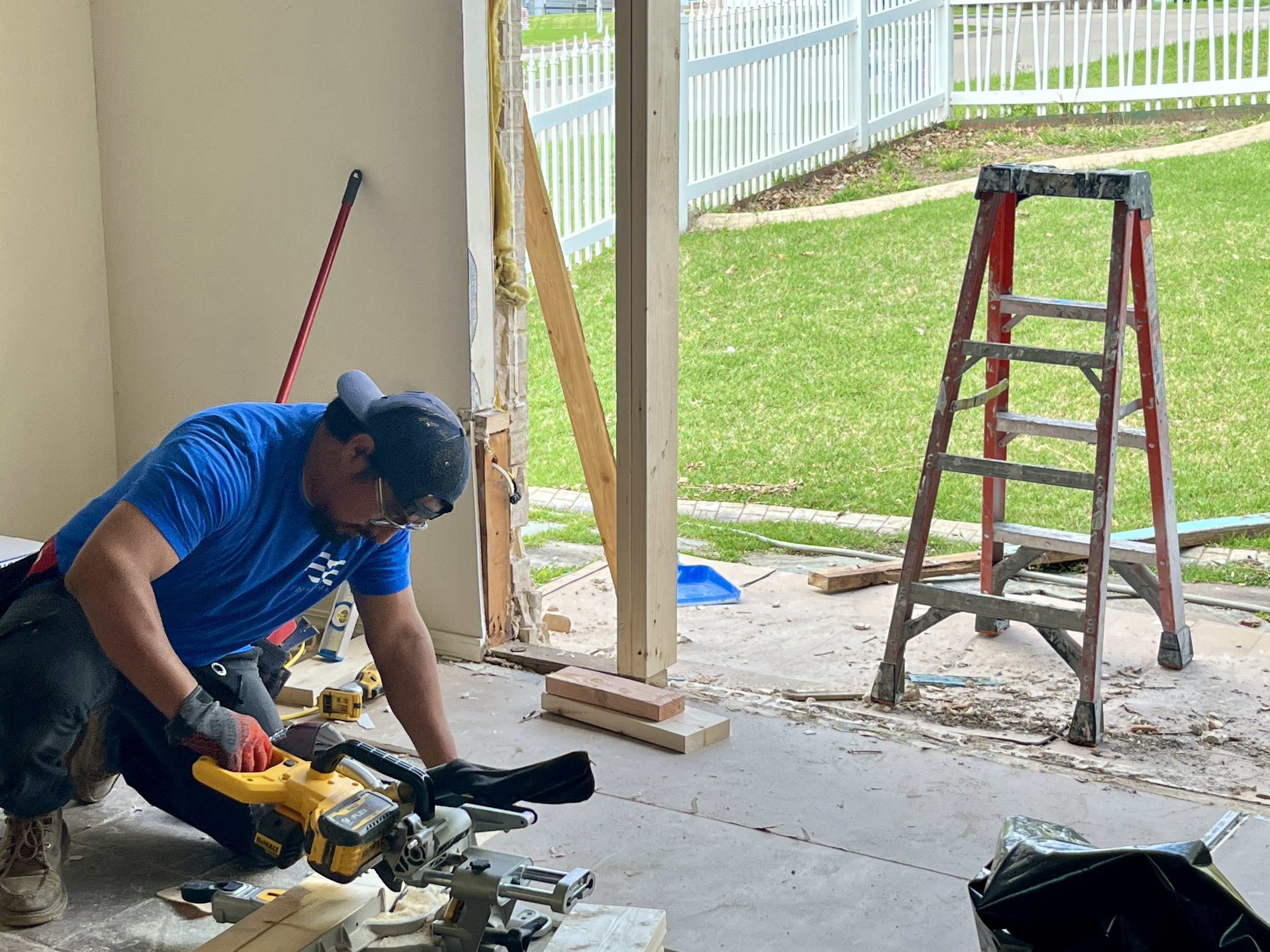 A construction worker wearing a hat and glasses is working inside on a house renovation project, using a power saw on a piece of wood. There is a red step ladder outside, and a grassy yard with a white fence in the background.