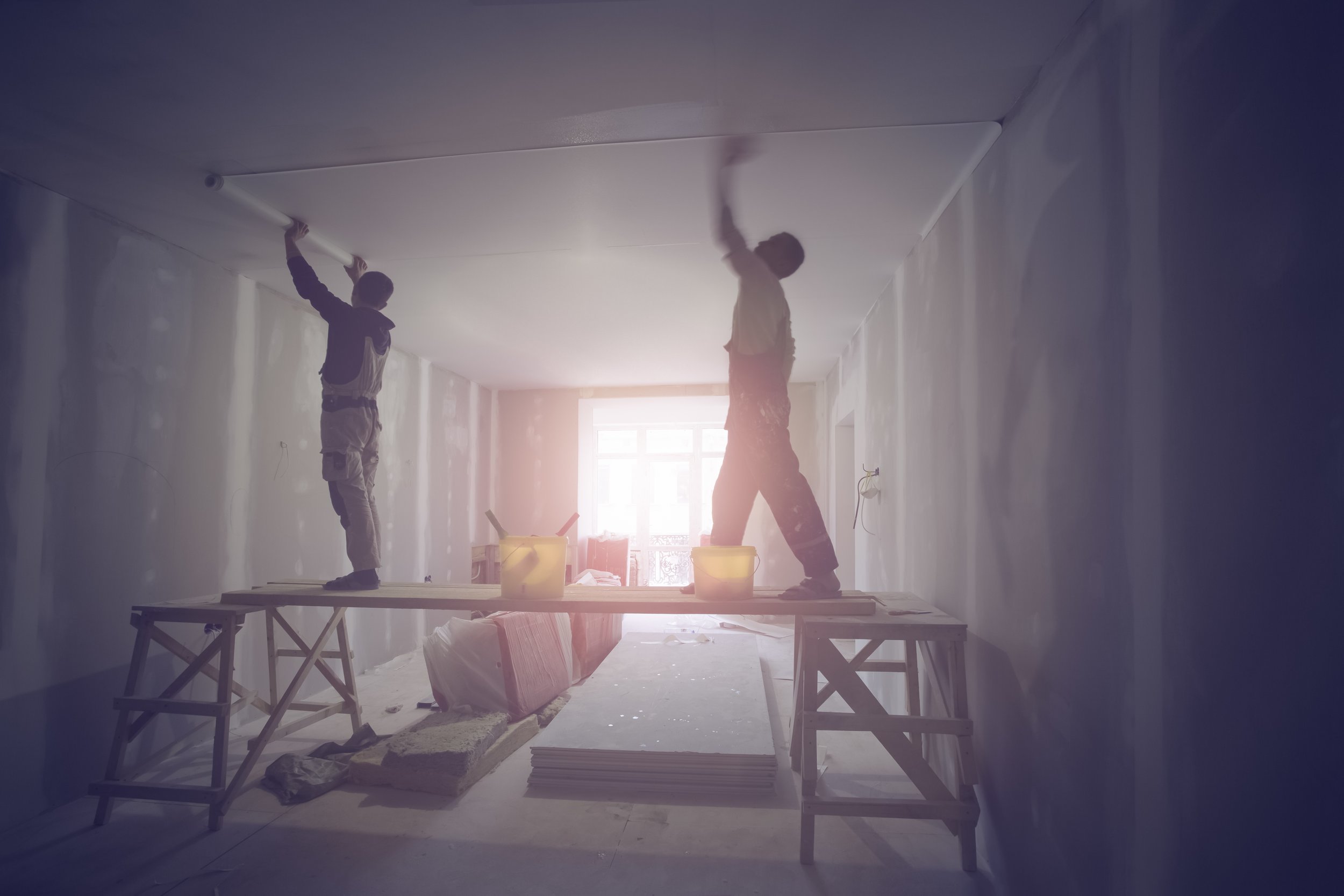 Two workers standing on a wooden plank supported by saw horses, installing or working on the ceiling of a room under construction. The room has unfinished walls and a large window providing natural light.