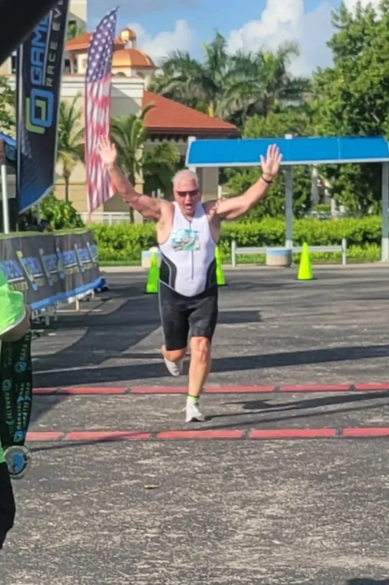 David Mark Decherd running a race, crossing the finish line with arms raised in victory, wearing sunglasses, a white sleeveless shirt, black shorts, and white running shoes, in an outdoor setting with trees, buildings, and safety cones visible in the