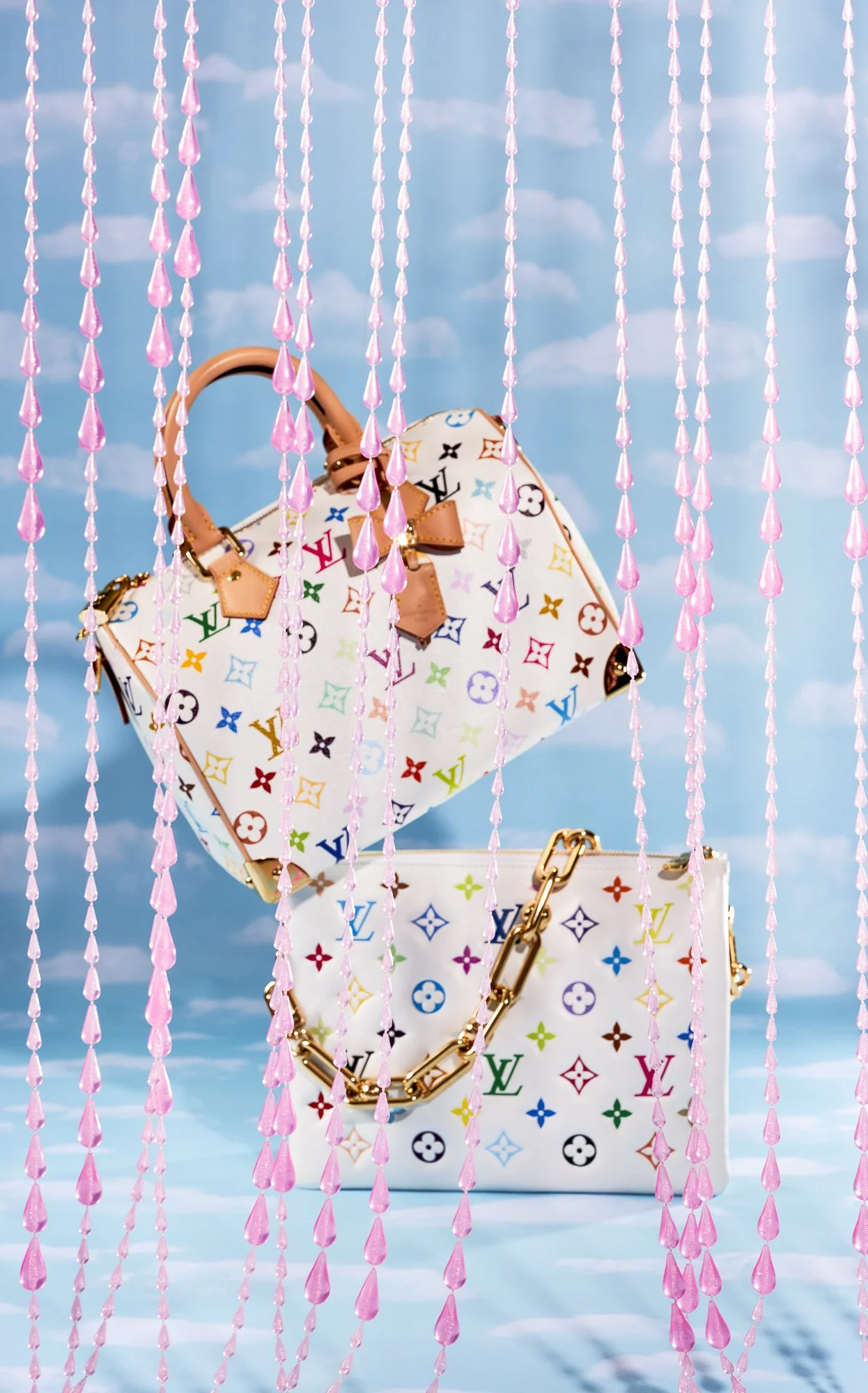 Two Louis Vuitton handbags with multicolored logos are seen behind pink hanging beads against a blue sky background.