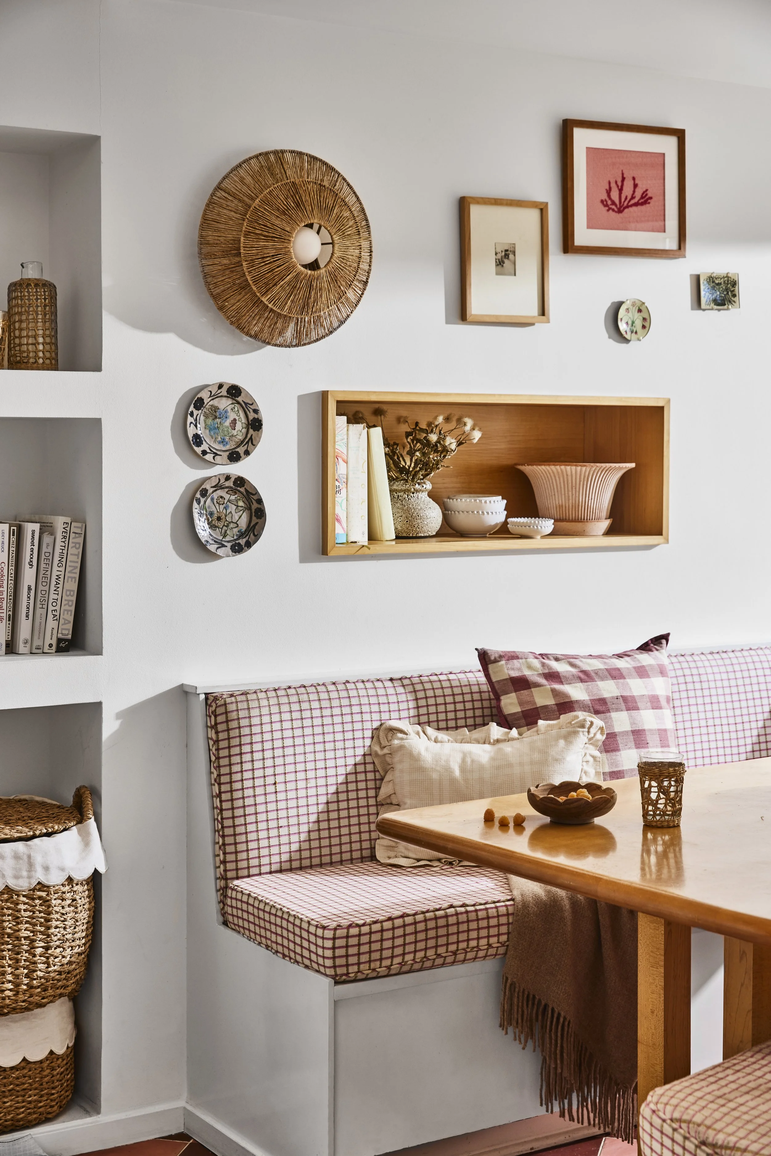 A cozy dining nook with a built-in bench featuring red and white checkered cushions, a wooden table with a bowl of snacks and a drink, and a white wall decorated with framed artwork, ceramic plates, and a woven wall hanging. There are also woven baskets on the side.