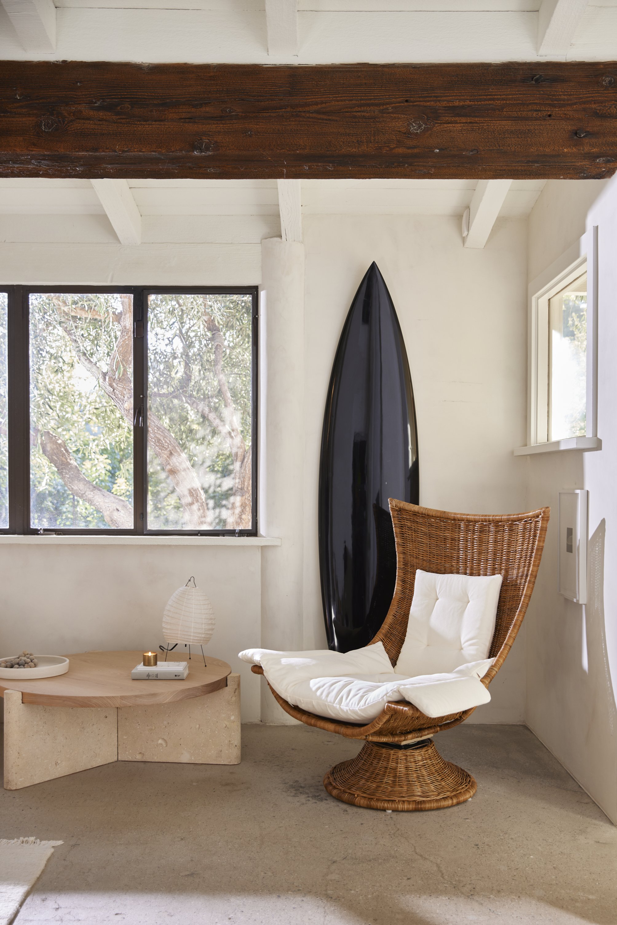 A cozy corner with a large window showing trees outside, a wicker chair with a white cushion, a black surfboard mounted on the wall, a small wooden table with a paper lantern and decorative bowl, and a textured beige carpet on the floor.