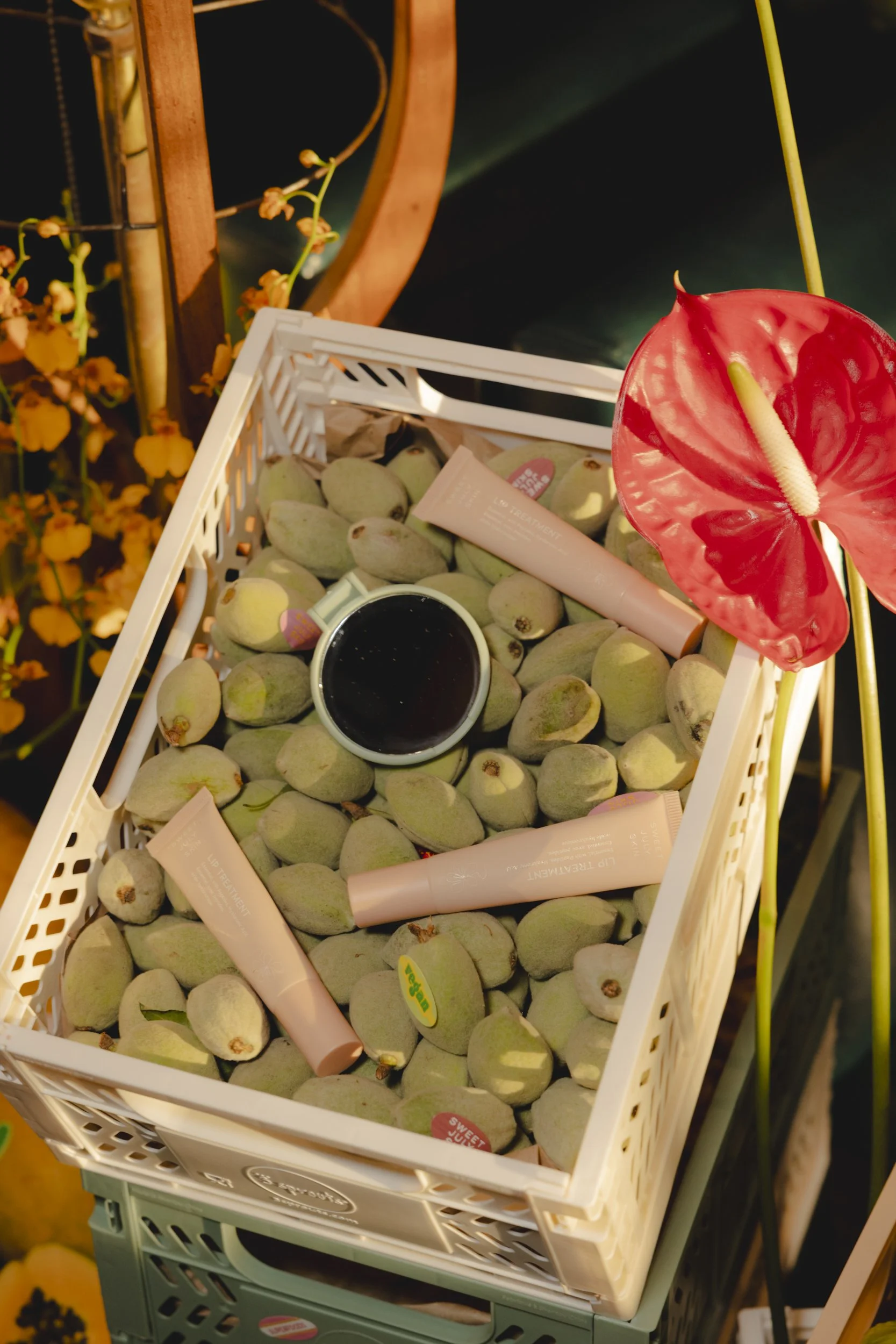 A white basket filled with green fruits, possibly figs, with a small cup of black coffee and pink skincare tubes on top. An anthurium flower with a red, heart-shaped leaf and white spadix is attached to the side of the basket.