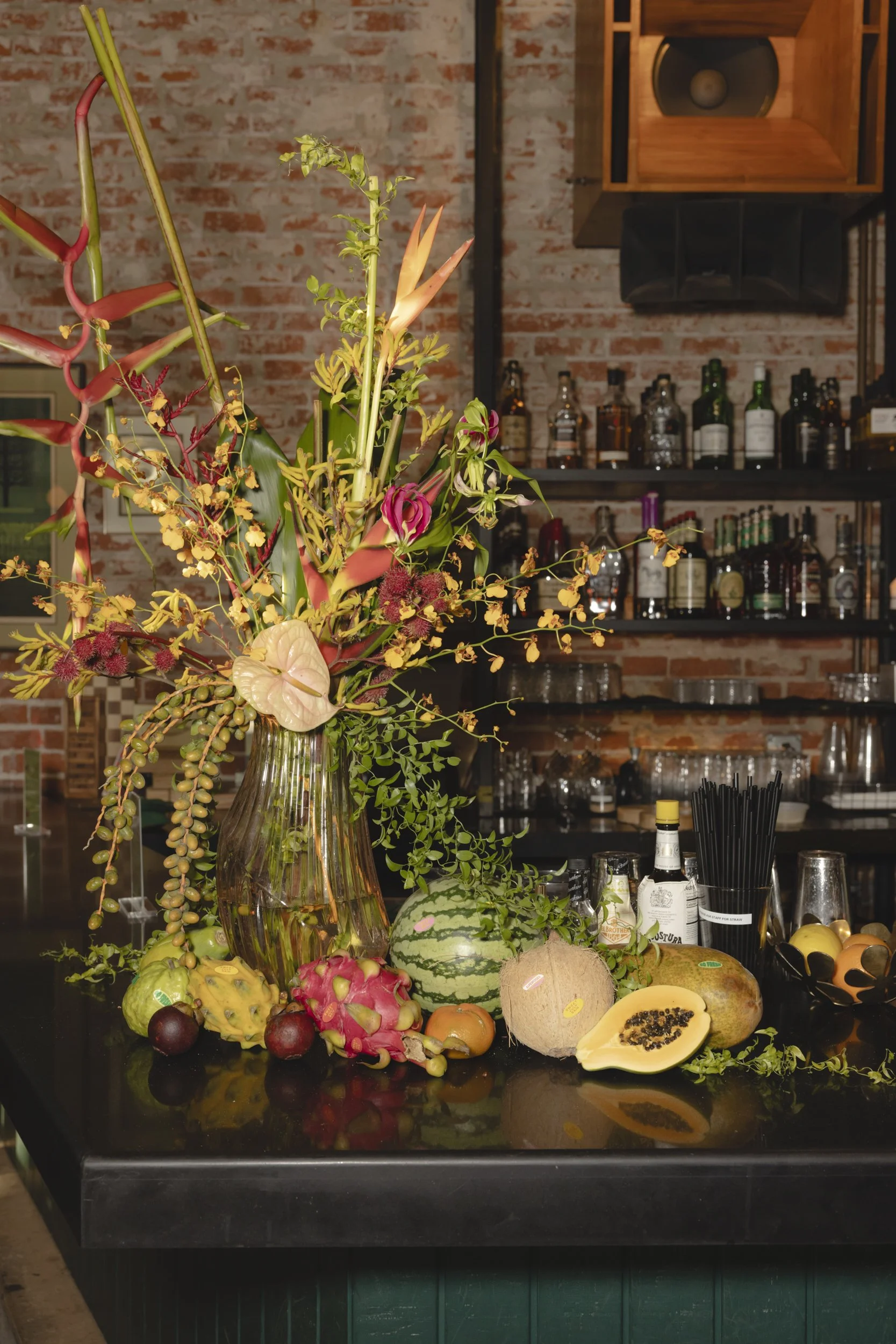 An elaborate floral arrangement with tropical fruits on a black countertop in a bar setting.