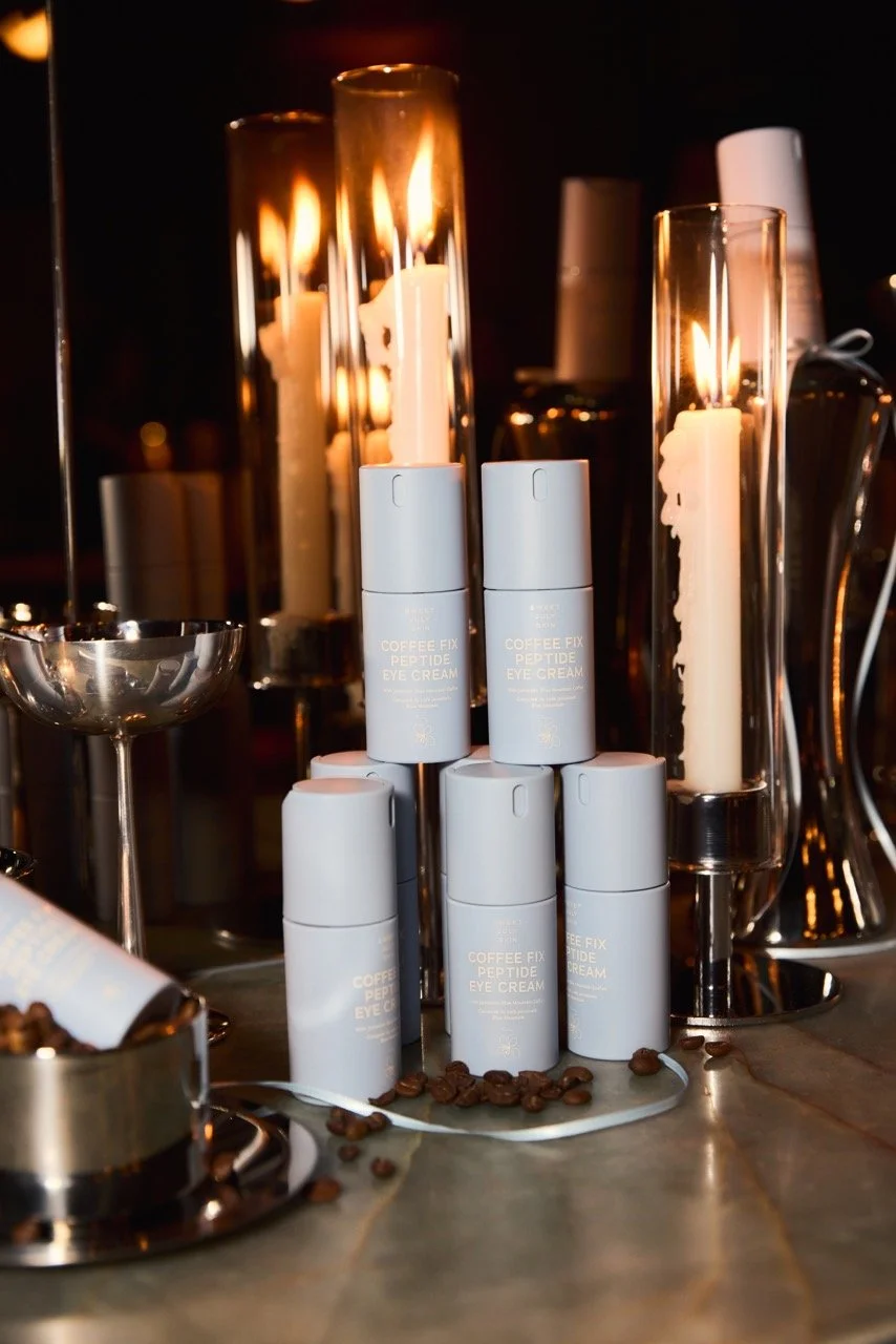 Stacked bottles of Coffee Fix Peptide Eye Cream surrounded by lit candles and scattered coffee beans on a table.