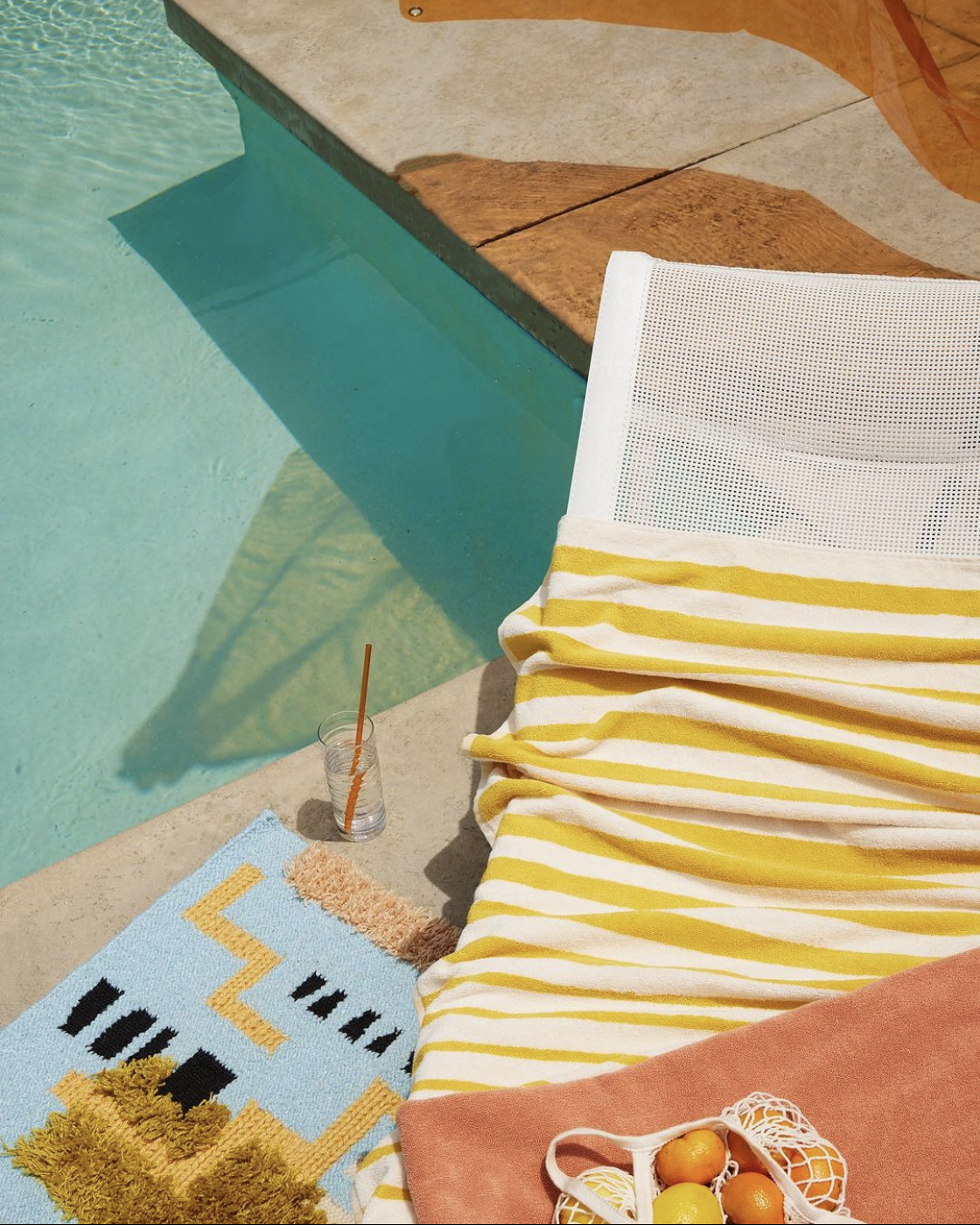 Poolside scene with a white and yellow striped towel on a lounge chair, a glass with an orange straw, a colorful rug with geometric designs, a small basket of fruit, and the pool with clear blue water in the background.