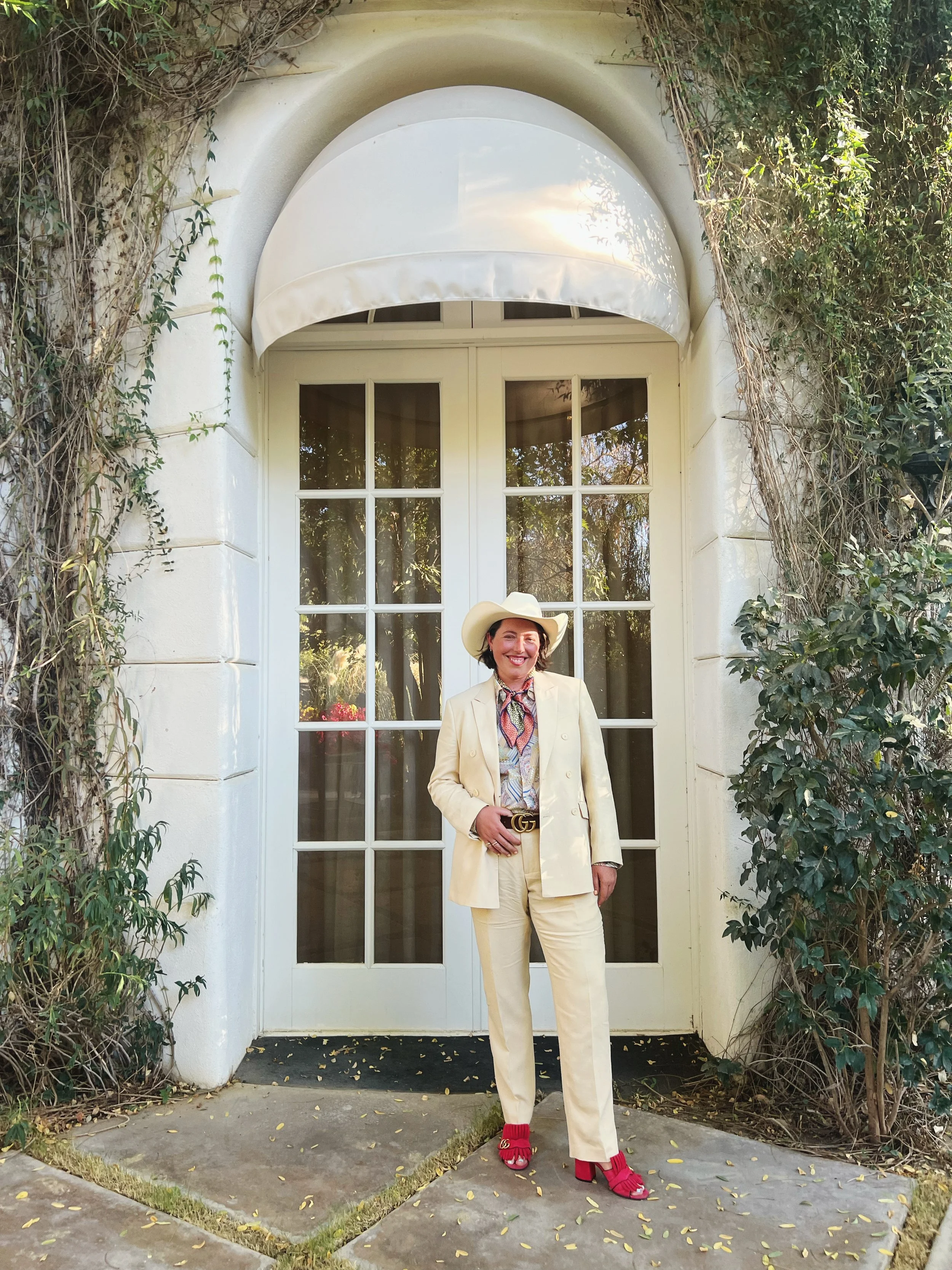 A non-binary person dressed in a cream suit, pink gucci heels, and a cowboy hat standing in front of a white door with glass panes, surrounded by greenery.