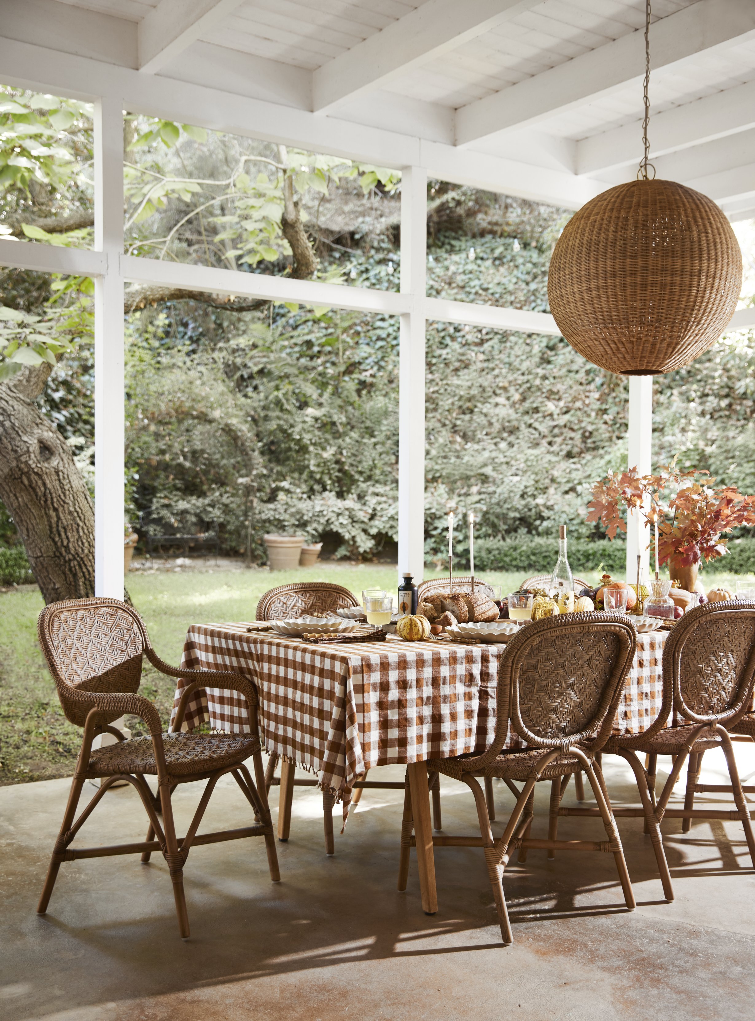 A table with a checked tablecloth set for a meal, on a screened porch with outdoor greenery, wicker chairs, and a large hanging light fixture.