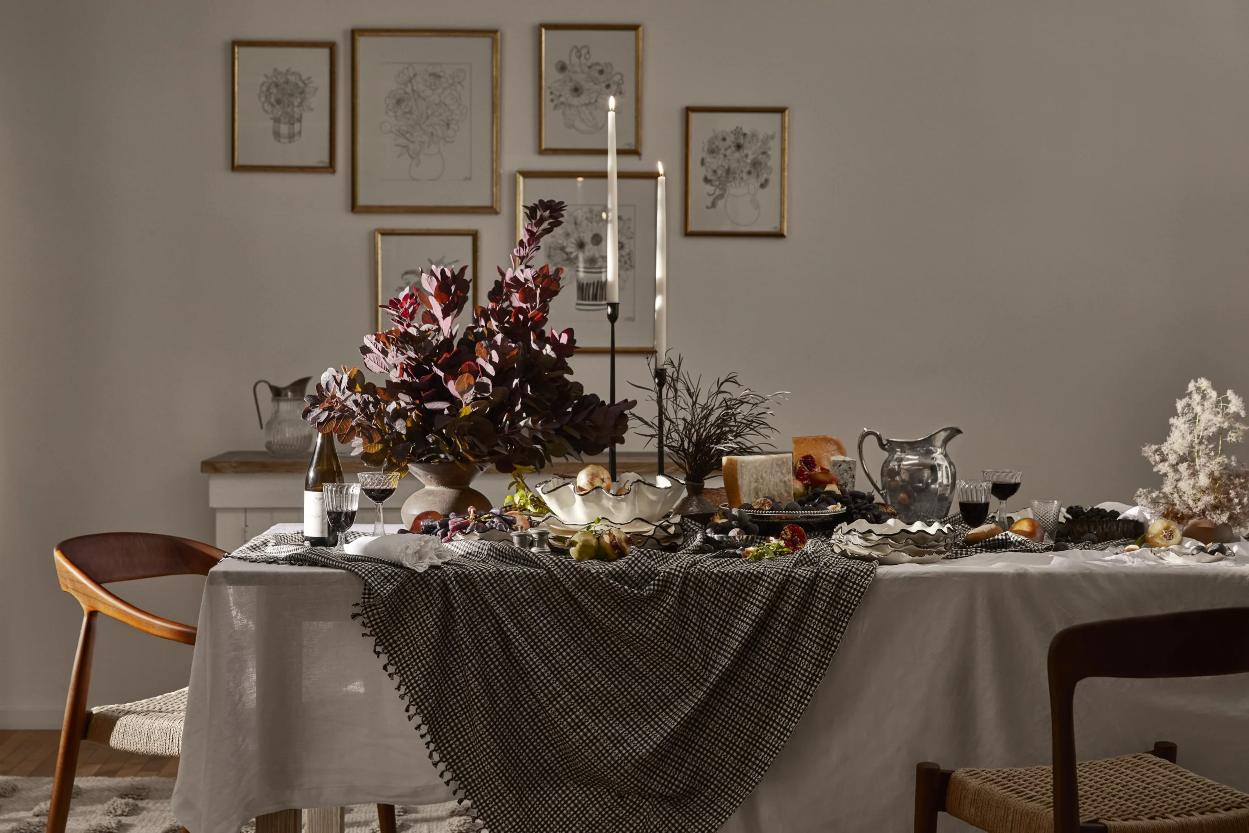 A dining table set for a meal with a white tablecloth, decorated with a large dark burgundy leafy centerpiece, candles, wine glasses, cheese, and fruit, with framed floral artwork on the wall behind.