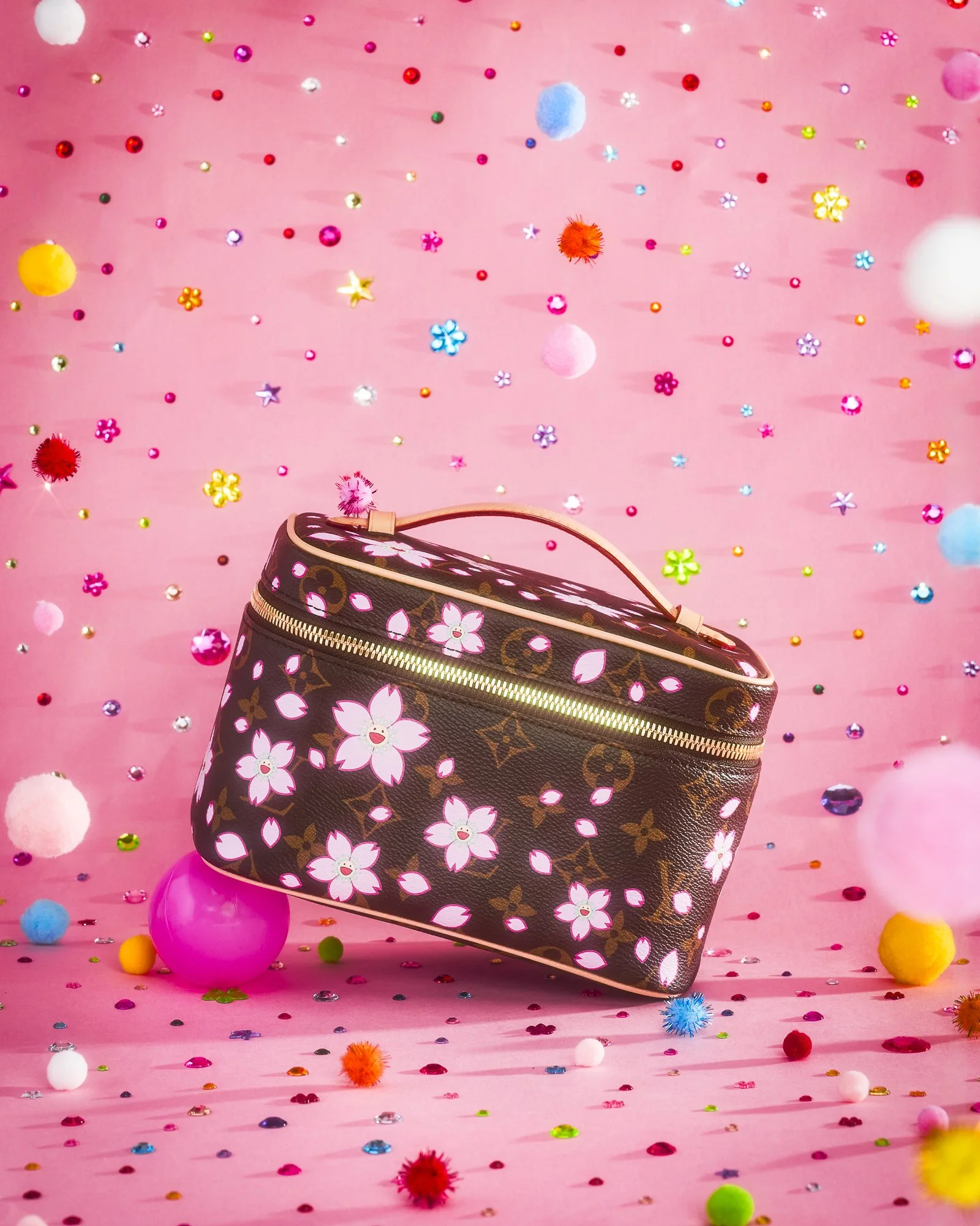 A decorated Louis Vuitton bag with floral patterns surrounded by colorful pom-poms, confetti, and small decorative objects on a pink background.