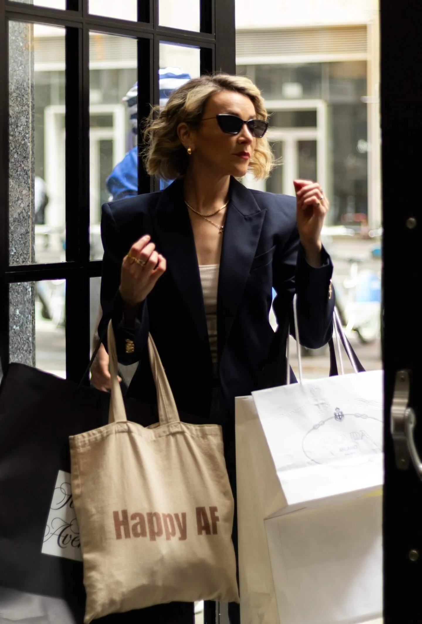 A woman wearing sunglasses, a dark blazer, and jewelry, carrying shopping bags, standing at a door while holding her fists up.