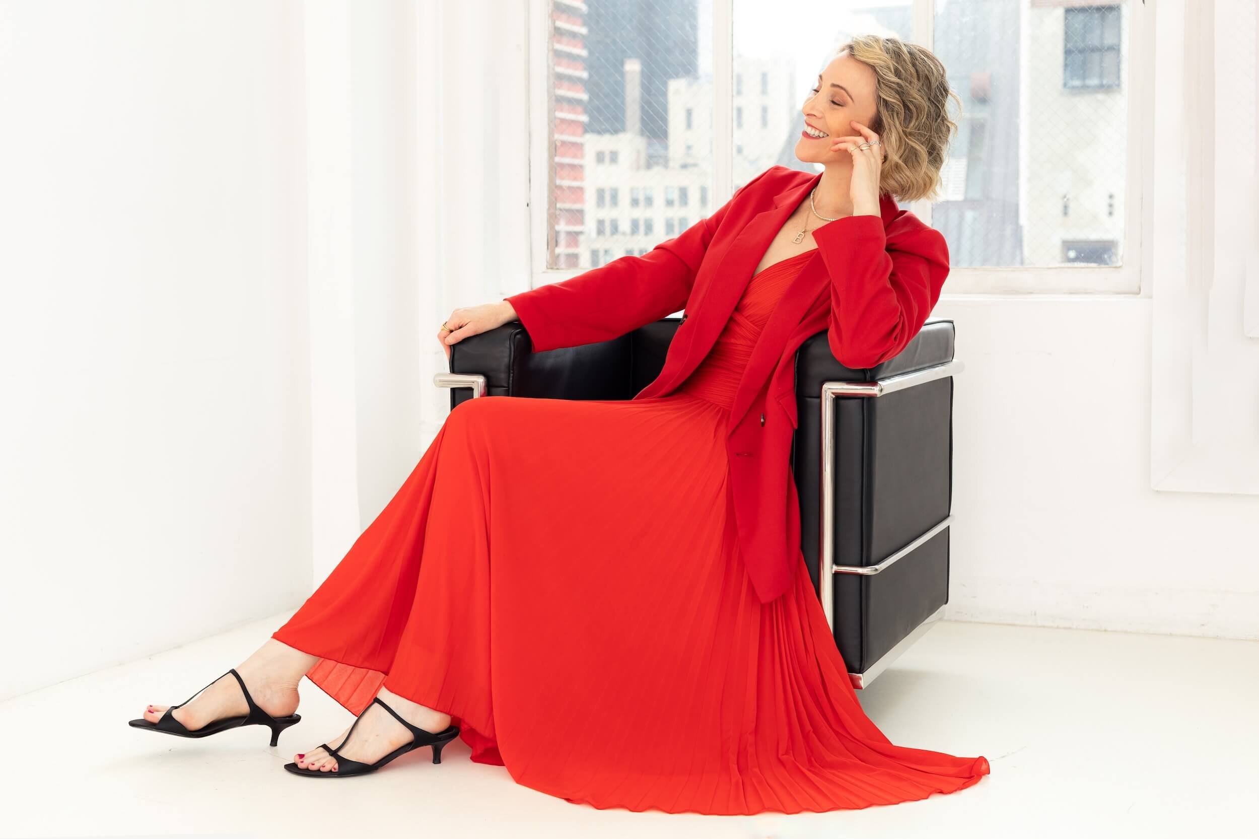 Woman sitting on a black chair near a window, wearing a long red dress and matching blazer, smiling with her eyes closed, with city buildings visible outside.