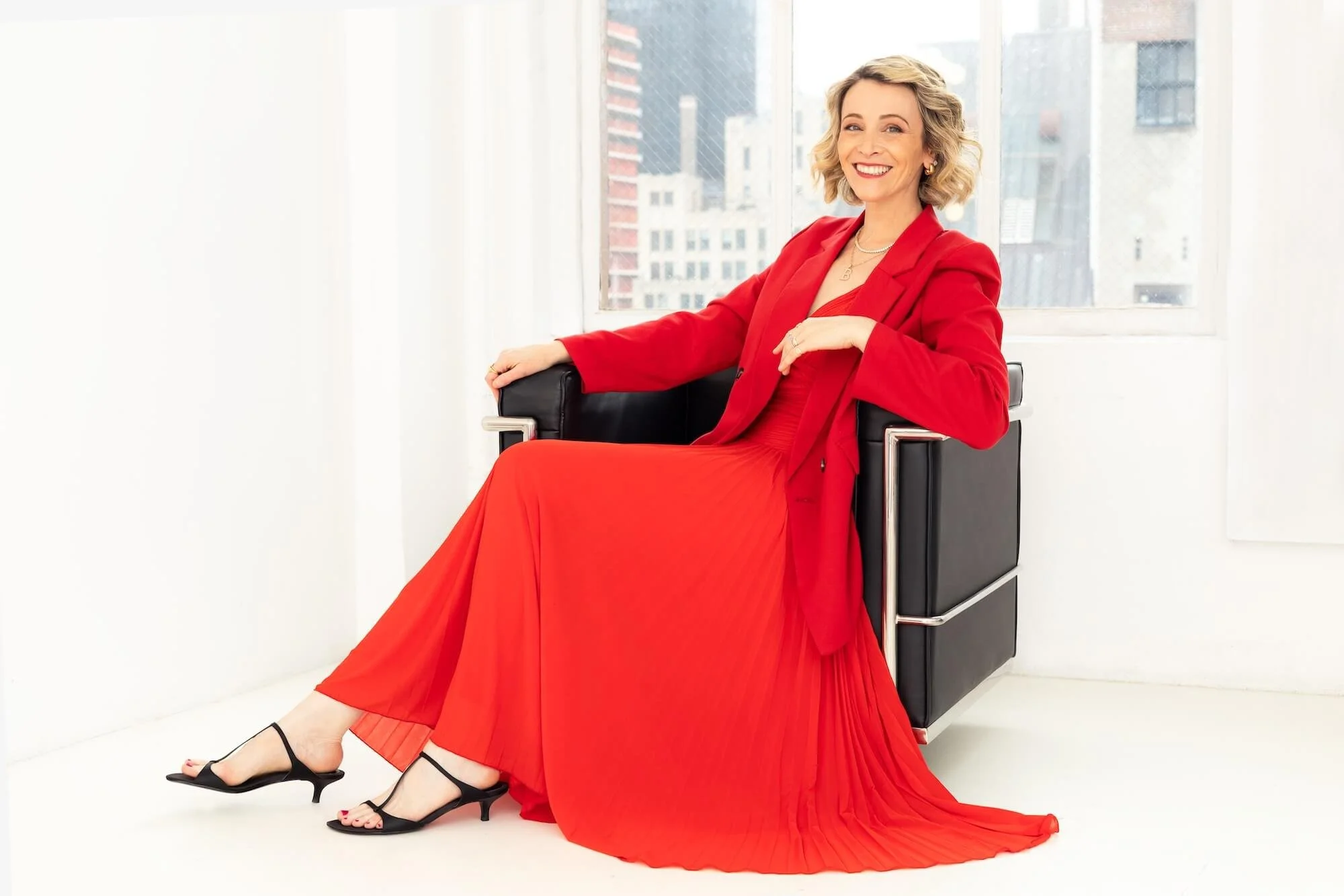 A woman in a red dress and blazer, with black high heels, sitting on a black chair in a bright room with large windows and cityscape background, smiling.