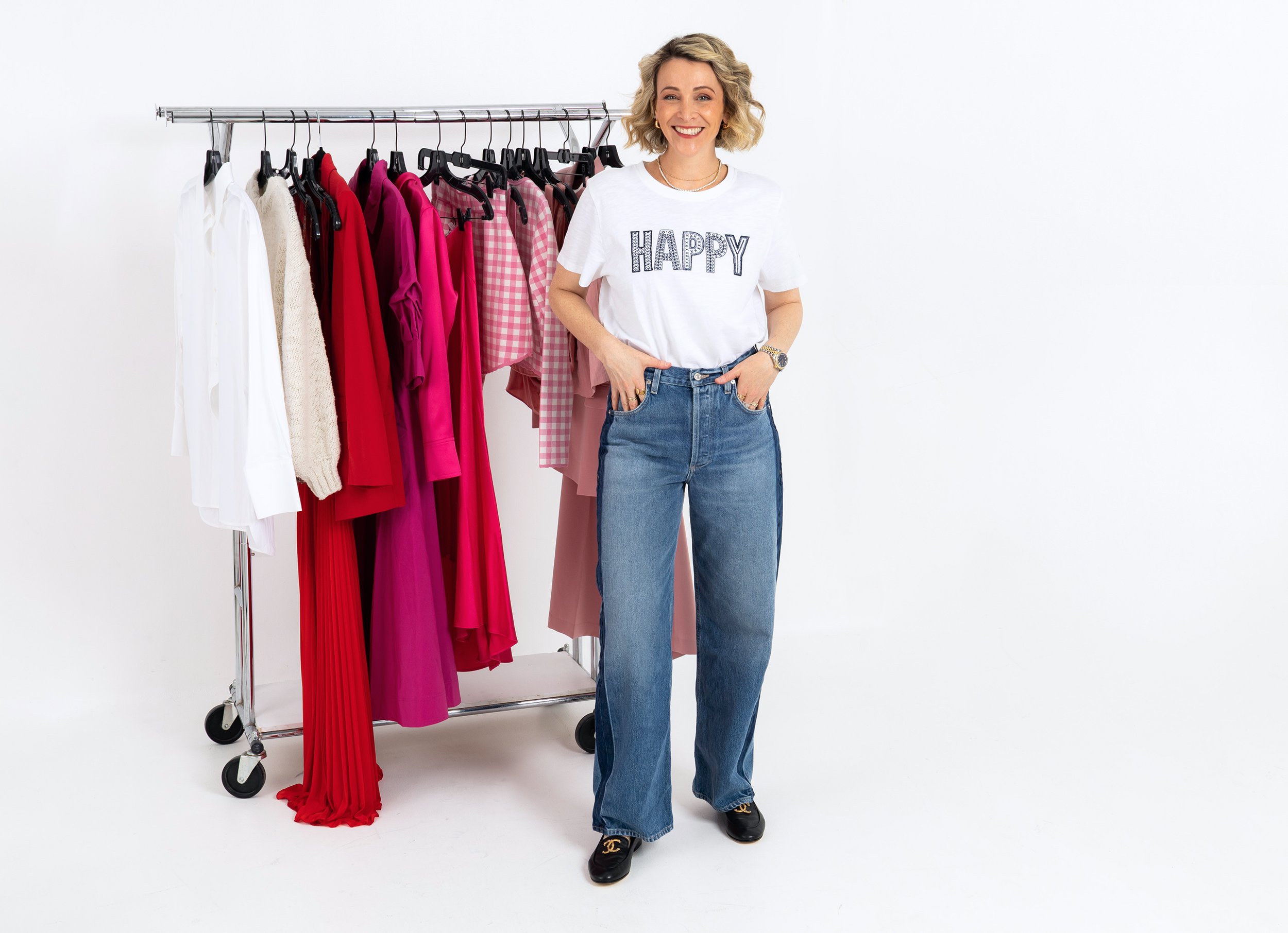 A woman with blonde wavy hair smiling, wearing a white t-shirt with 'HAPPY' written on it and blue jeans, stands next to a clothing rack with various pink, red, and white clothing items against a white background.
