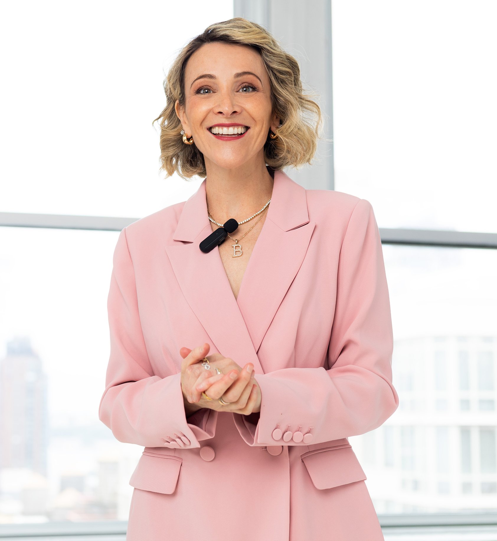 Woman with blonde curly hair smiling, wearing a pink blazer and jewelry, standing in front of a window with a cityscape view.
