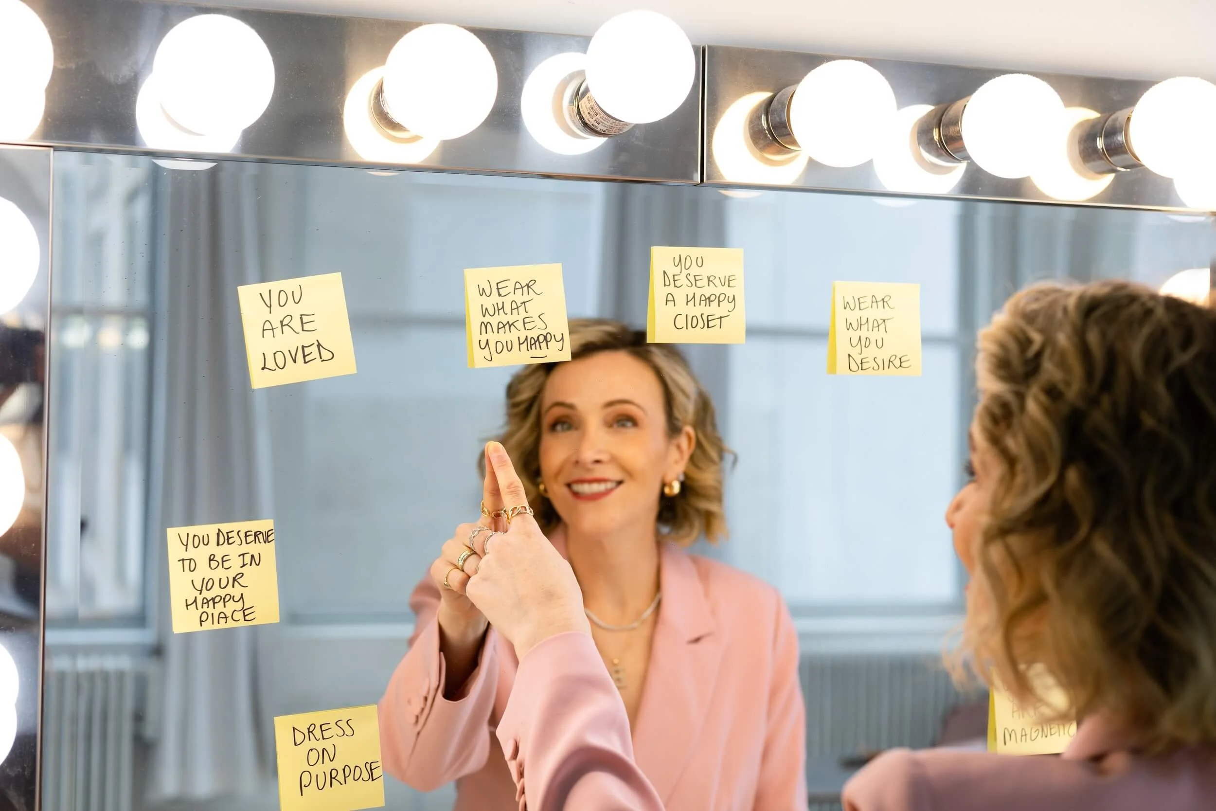 A woman smiling at her reflection in a dressing room mirror, surrounded by glowing vanity lights. She has various motivational sticky notes on the mirror, including phrases like 'You are loved,' 'Wear what makes you happy,' and 'Dress on purpose.' She is wearing a light pink blazer and gold jewelry.