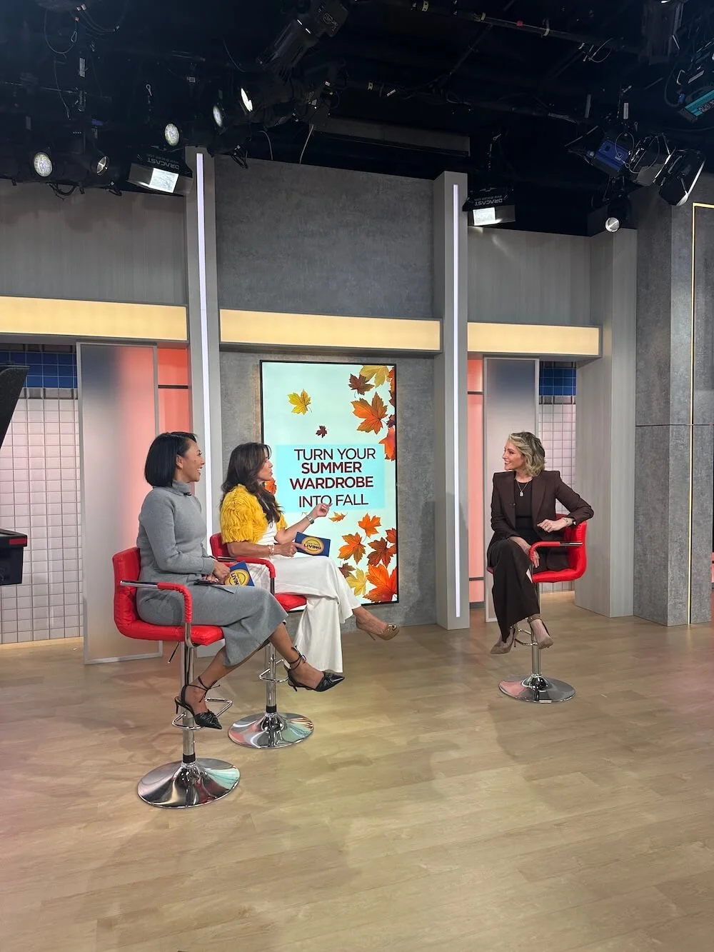 A daytime television set featuring three women seated on red chairs, discussing fall fashion with a large digital screen displaying the message "Turn Your Summer Wardrobe Into Fall" and autumn leaves background.