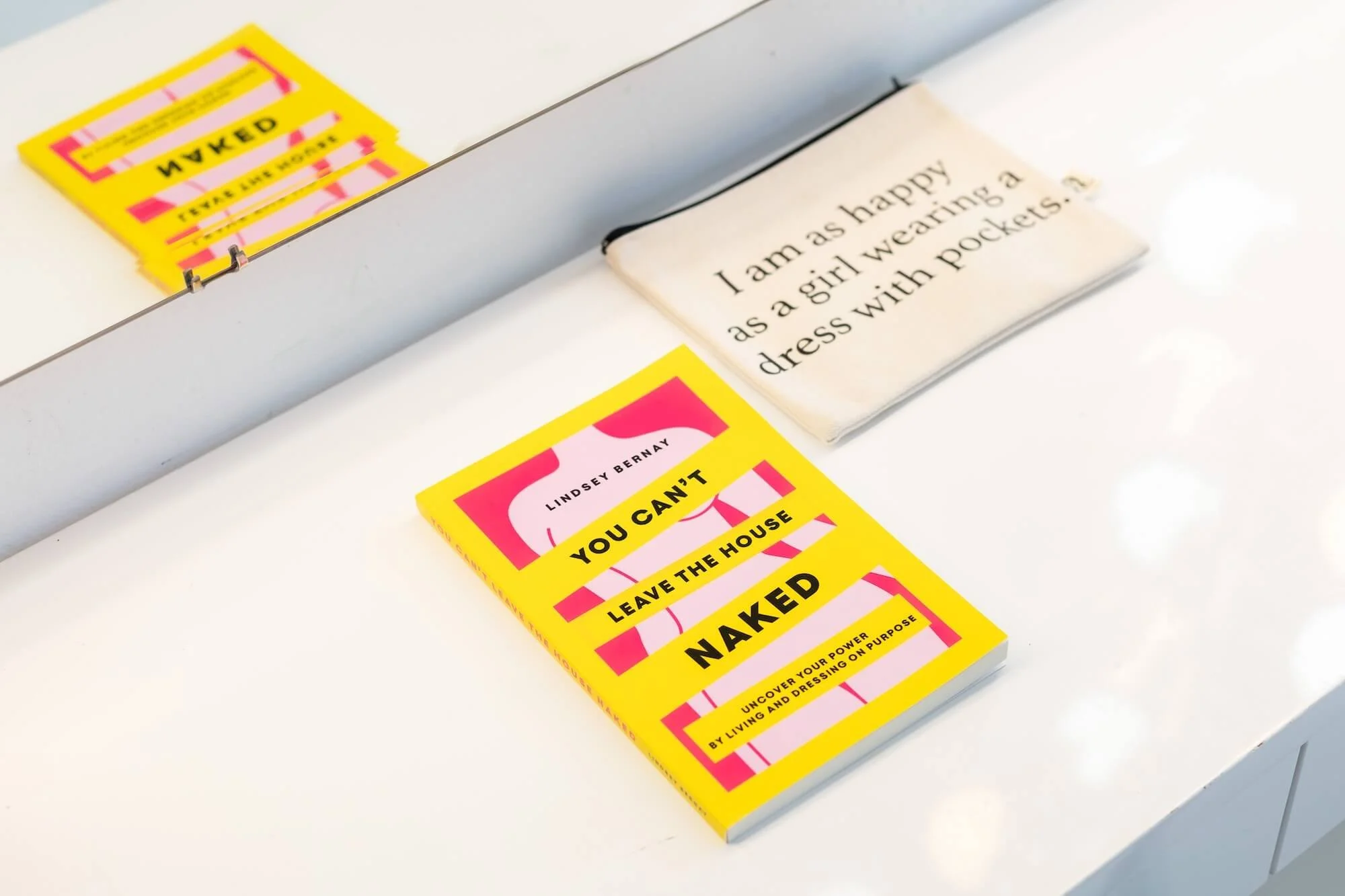 A yellow book titled 'You Can't Leave The House Naked' by Lindsey Bernay is on a white surface, with a mirror reflecting the book and a fabric item with text.