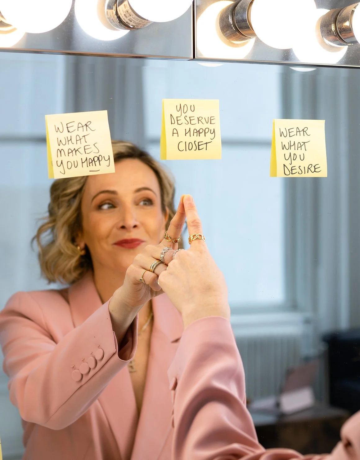 Woman in pink blazer reaching for yellow sticky notes on mirror, with motivational messages about happiness and fashion.