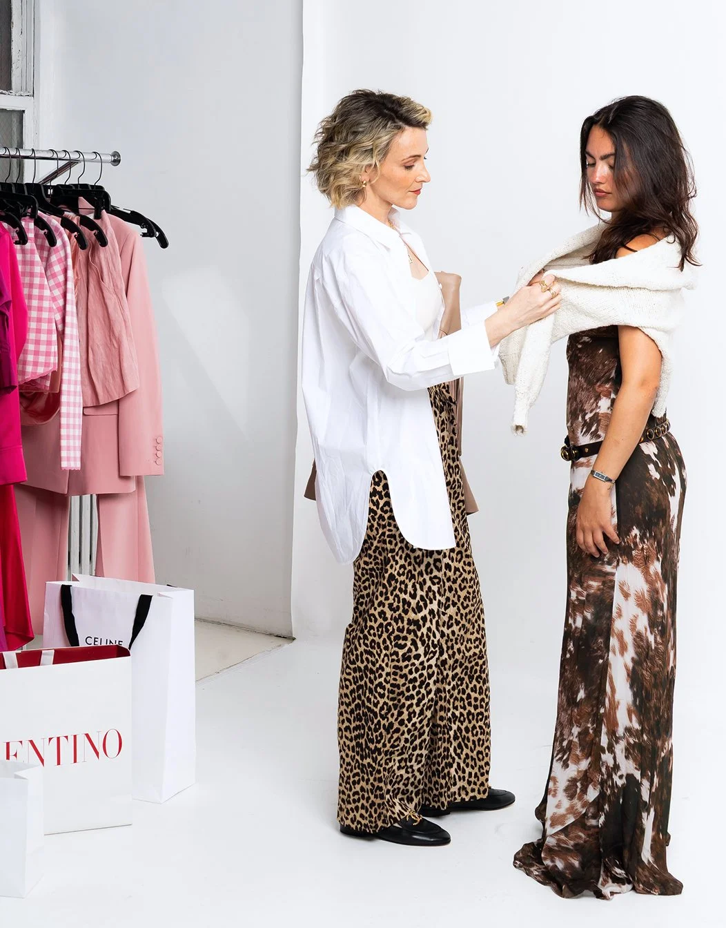 A woman with curly hair tries on a white sweater for a customer in a clothing store.