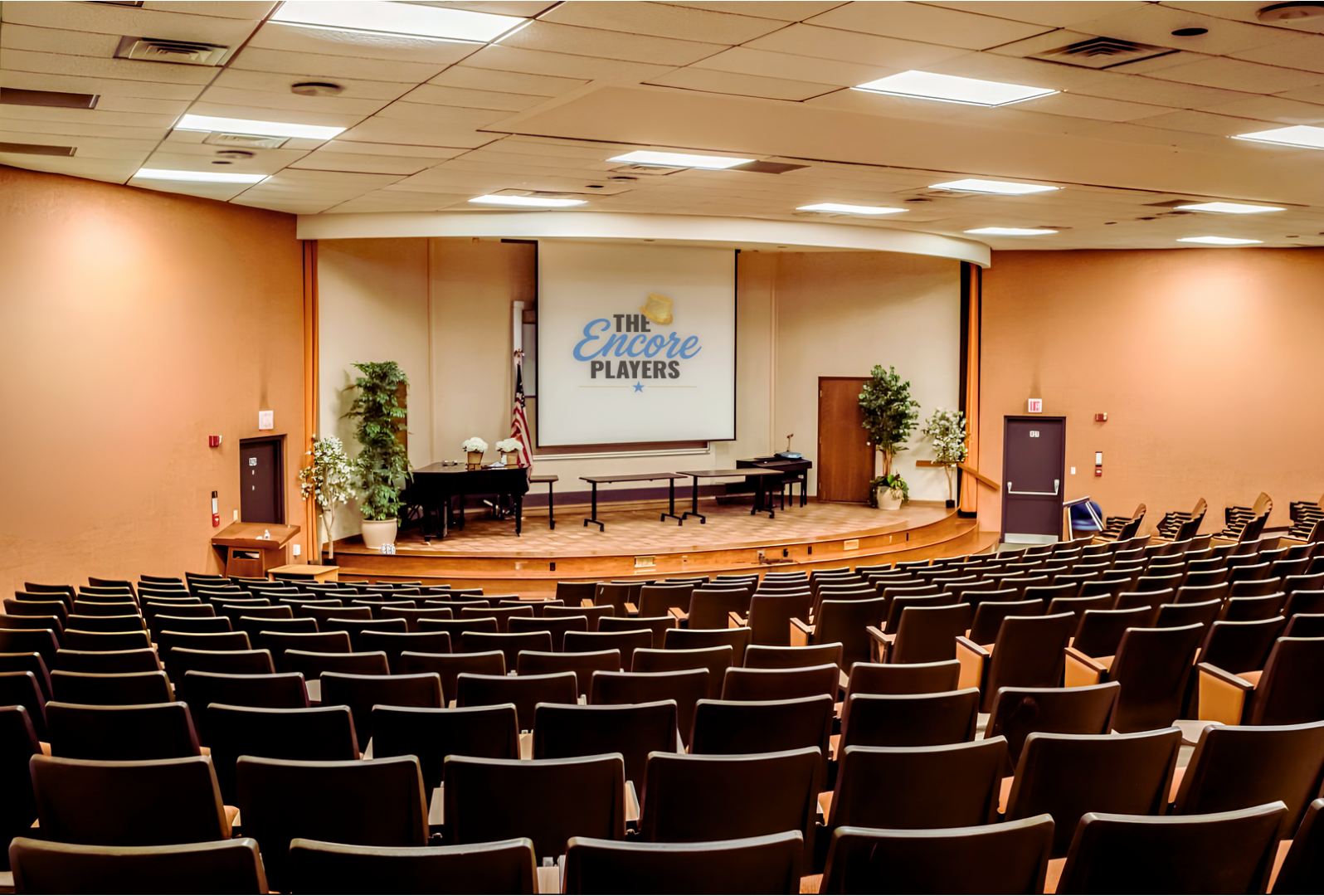 Empty auditorium with a stage, chairs facing the stage, a large screen displaying 'The Encore Players' and a logo, decorated with potted plants and flowers.