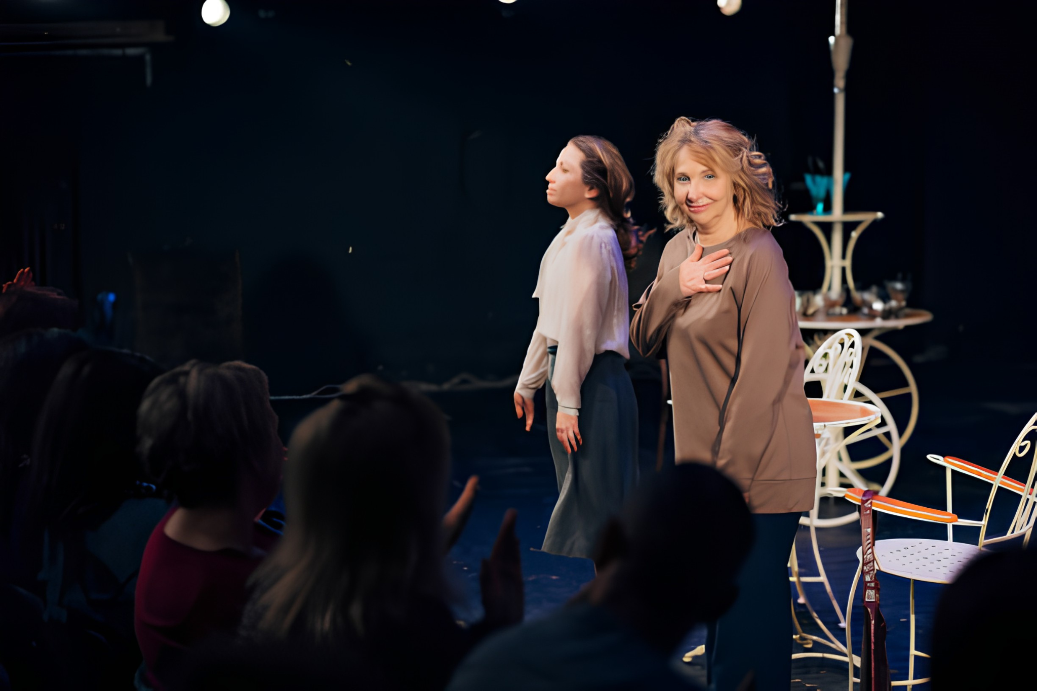 A woman with blonde hair smiling and placing her hand on her chest, standing next to a mannequin on stage. The woman is wearing a silk brown top. Behind them, there is a table with shoes and a vintage-style white bicycle. The audience is seated in the dark in front of the stage.