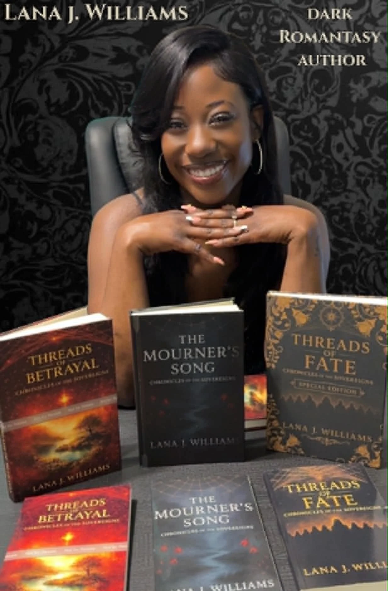A woman smiling, sitting behind a table with three books titled 'Threads of Betrayal,' 'The Mourner's Song,' and 'Threads of Fate' by Lana J. Williams. The background features a black ornate wall. All three books are displayed upright on the table, with additional copies in front of them.