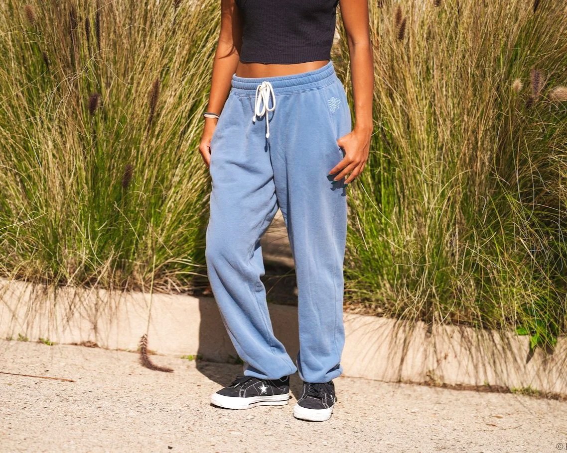 Photo of a person standing in front of tall grass - shown from the feet to the torso. Person is wearing light blue sweat pants and a black crop top.