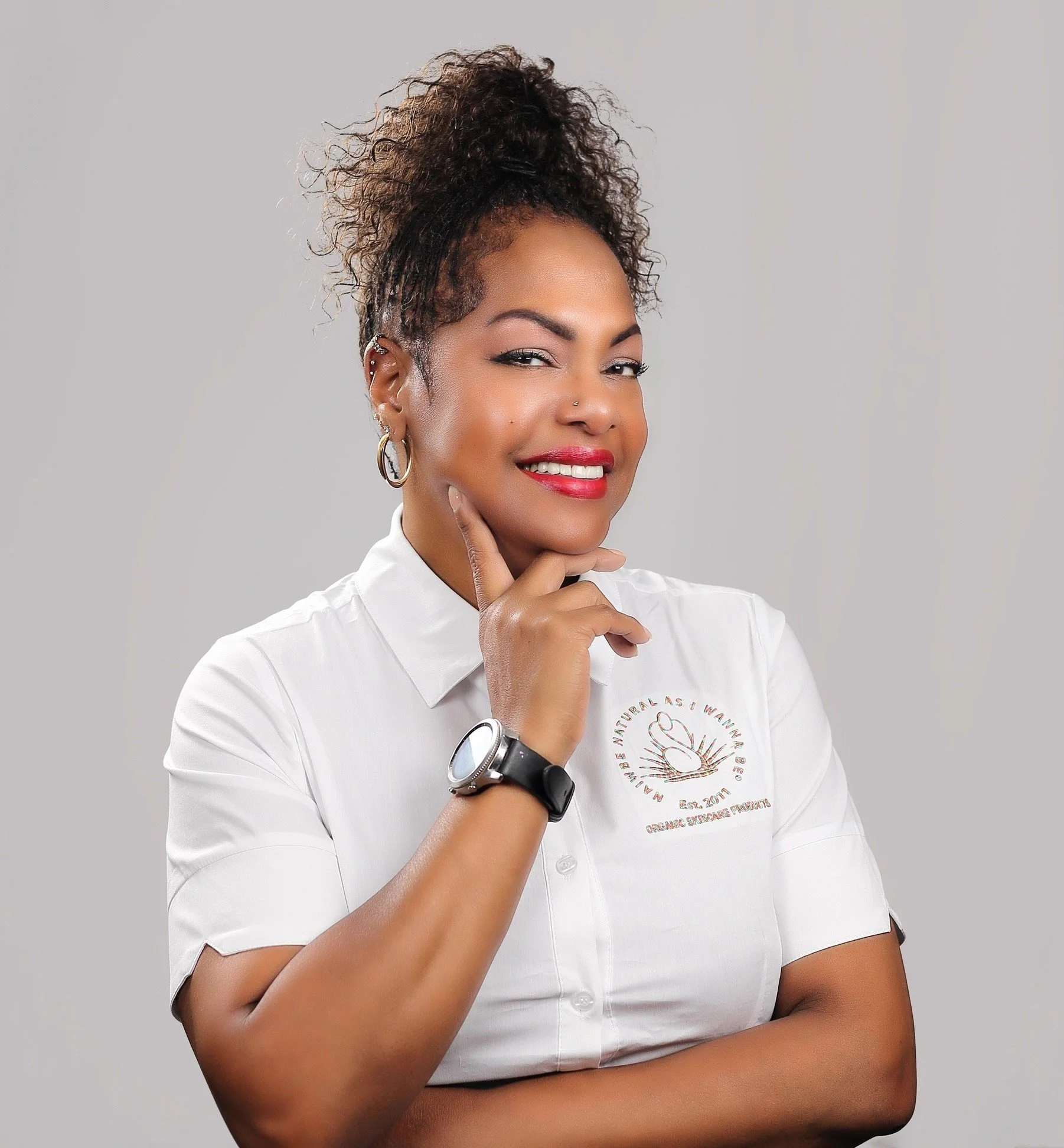 A woman with curly hair in a bun, wearing a white shirt and jewelry, smiling confidently, with one hand on her chin, against a plain gray background.
