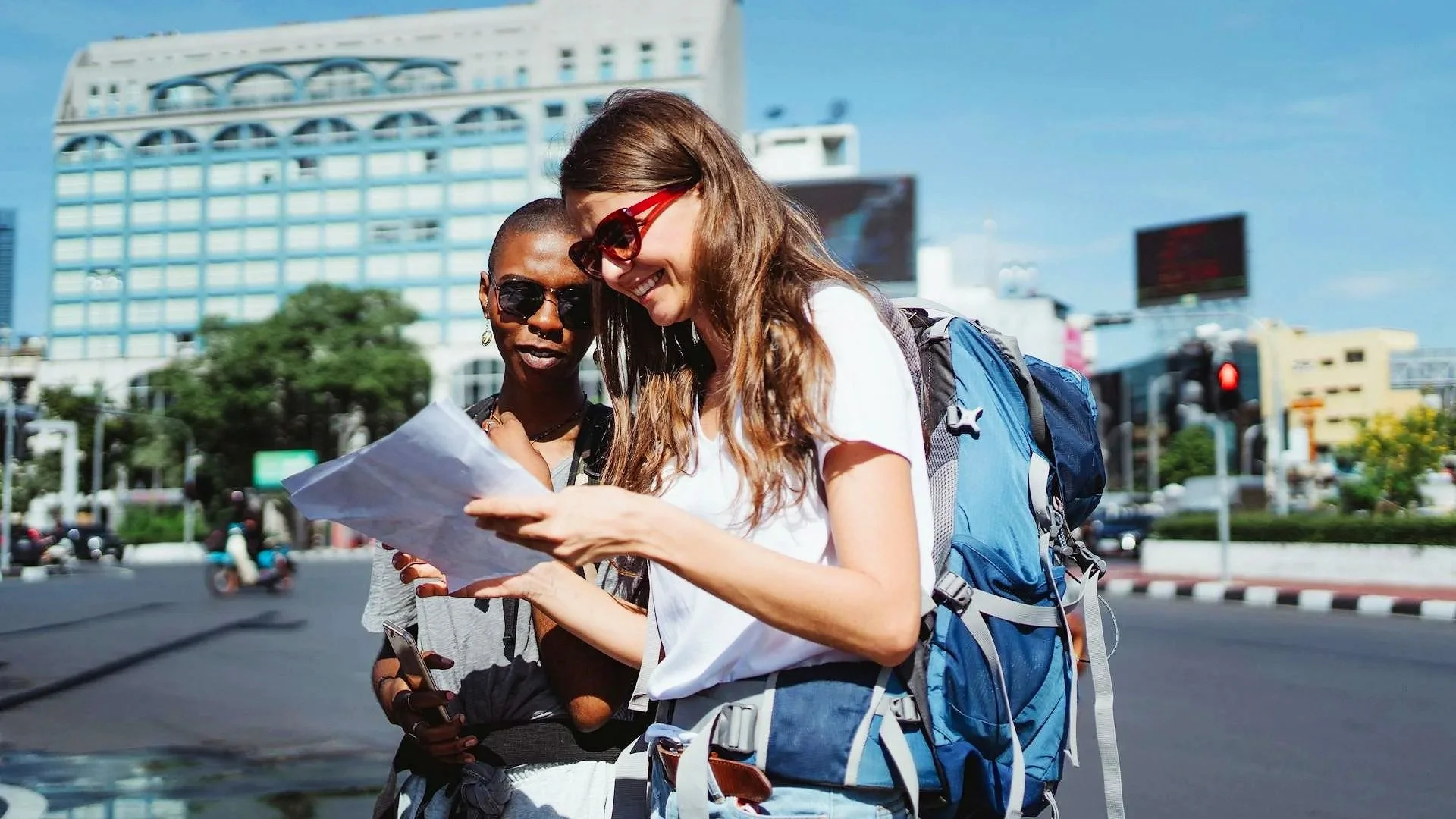A Black woman with a shaved head and a white woman with brunette hair stand side by side in a metropolitan area looking at some paper. They have travel backpacks on and sunglasses.