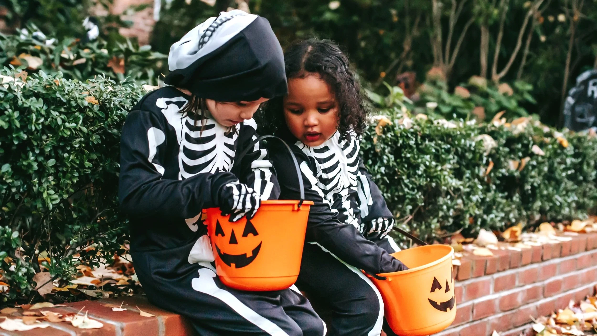 Two kids sit on a low brick wall, dressed as skeletons and looking into pumpkin trick or treat buckets