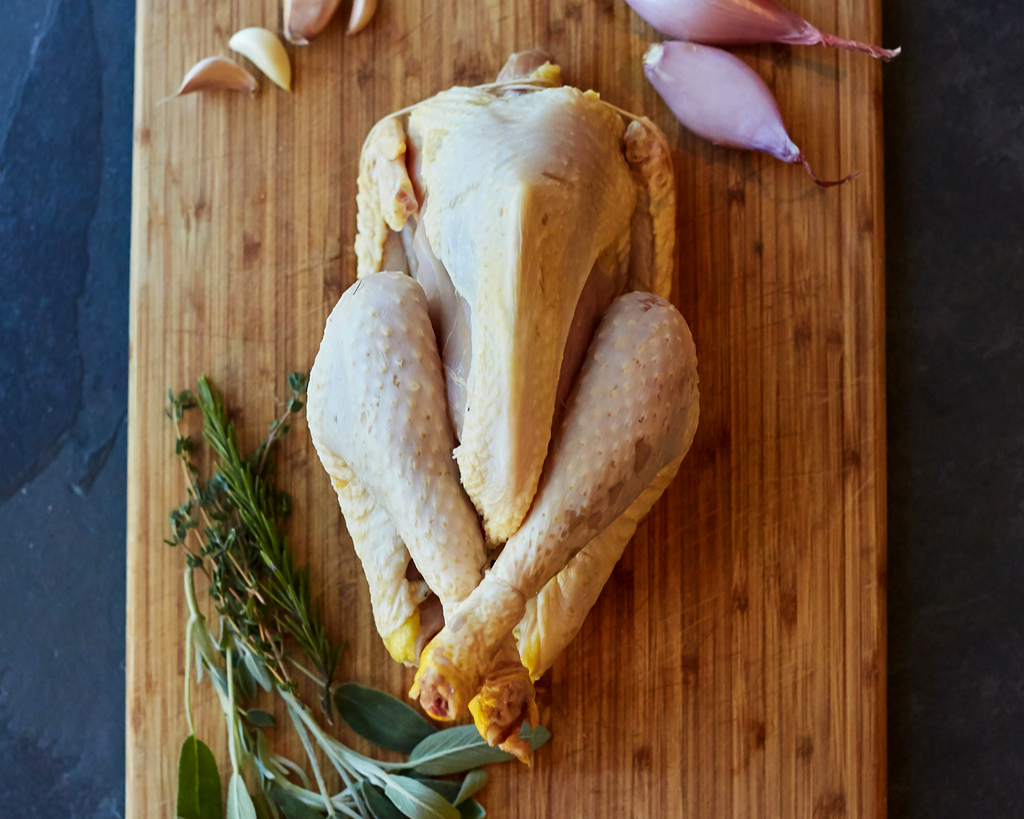 A raw whole chicken on a cutting board with some herbs at the bottom and garlic and shallots at the top