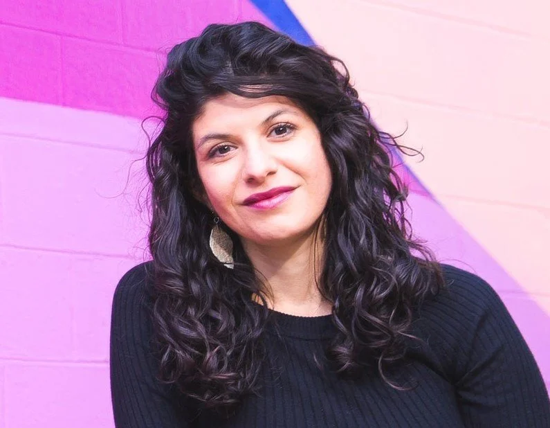 A Latina woman with long, dark curly hair in a black sweater poses before a brick wall painted various shades of pink