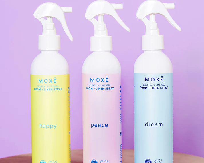 Three spray bottles against a lilac background. The bottles are white with different colored labels: yellow, light pink, light blue.