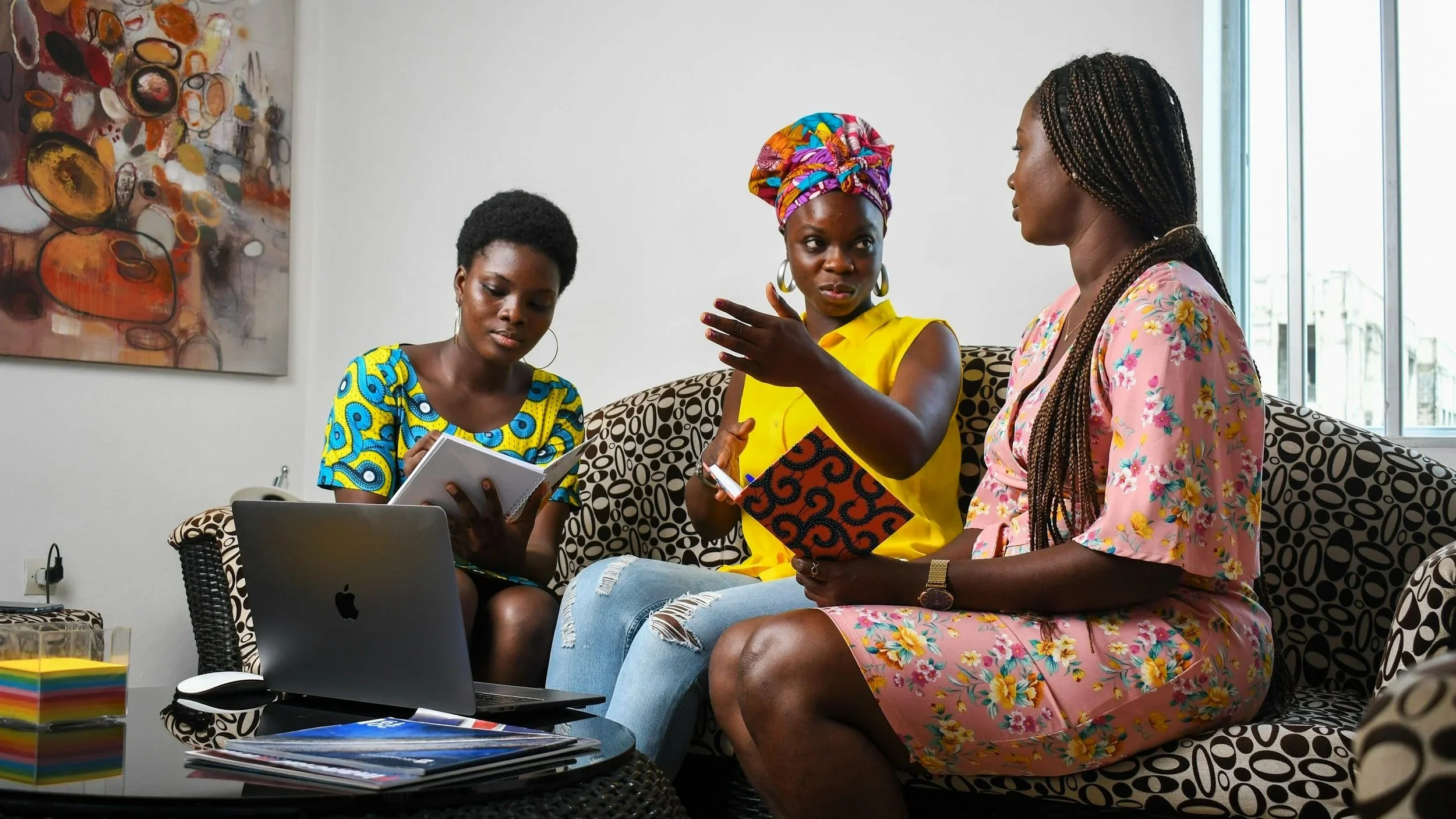 Three Black women sitting on a couch with a laptop in front of them and paperwork, in deep conversation