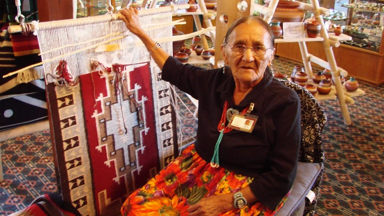 An older, Navajo woman sits at a weaving loom in a small store.