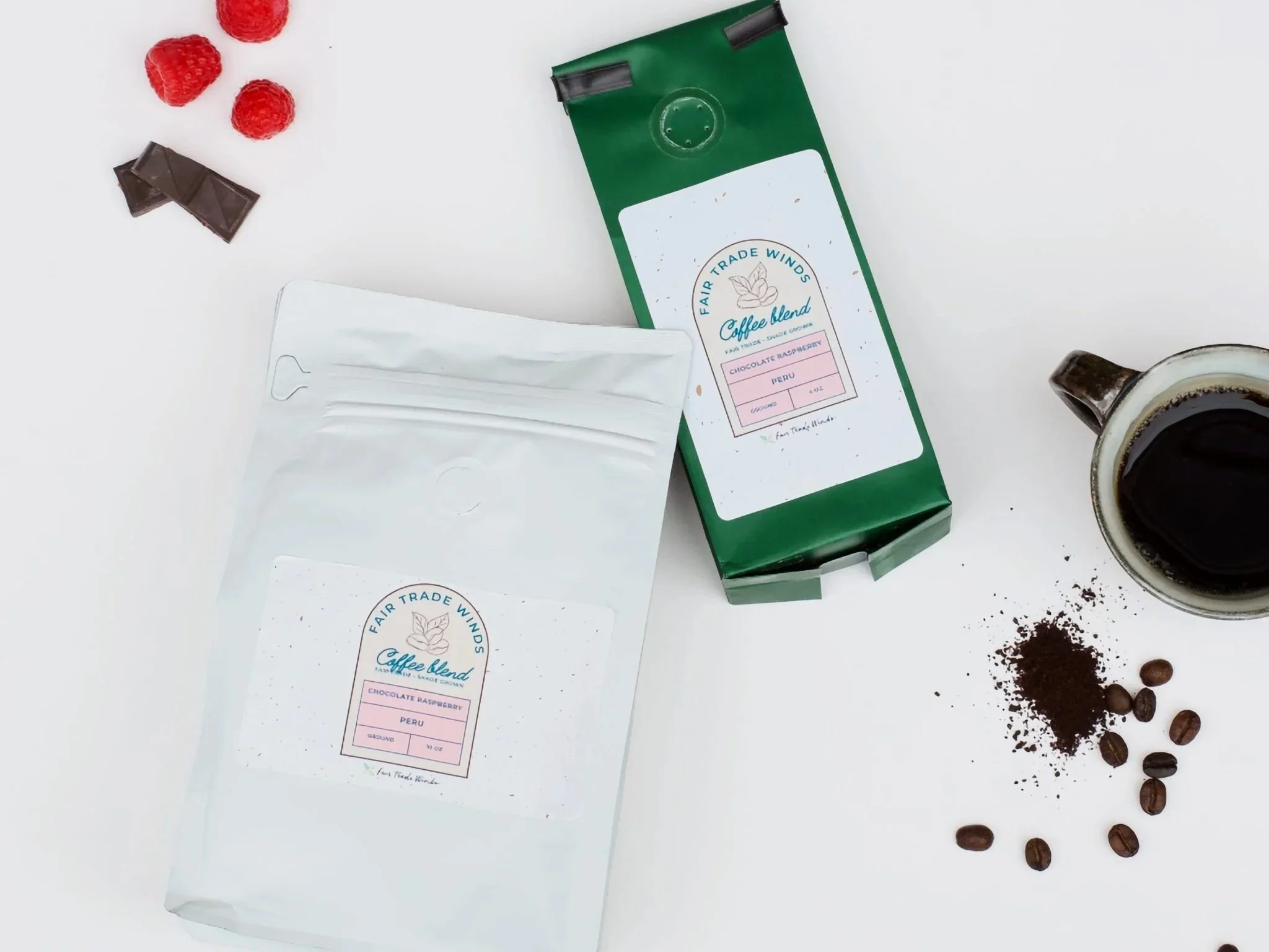 Two bags of coffee, one white and one green, sit on a white background with chocolate, raspberries, coffee beans, and a mug of coffee surrounding them, the angle is from above looking down
