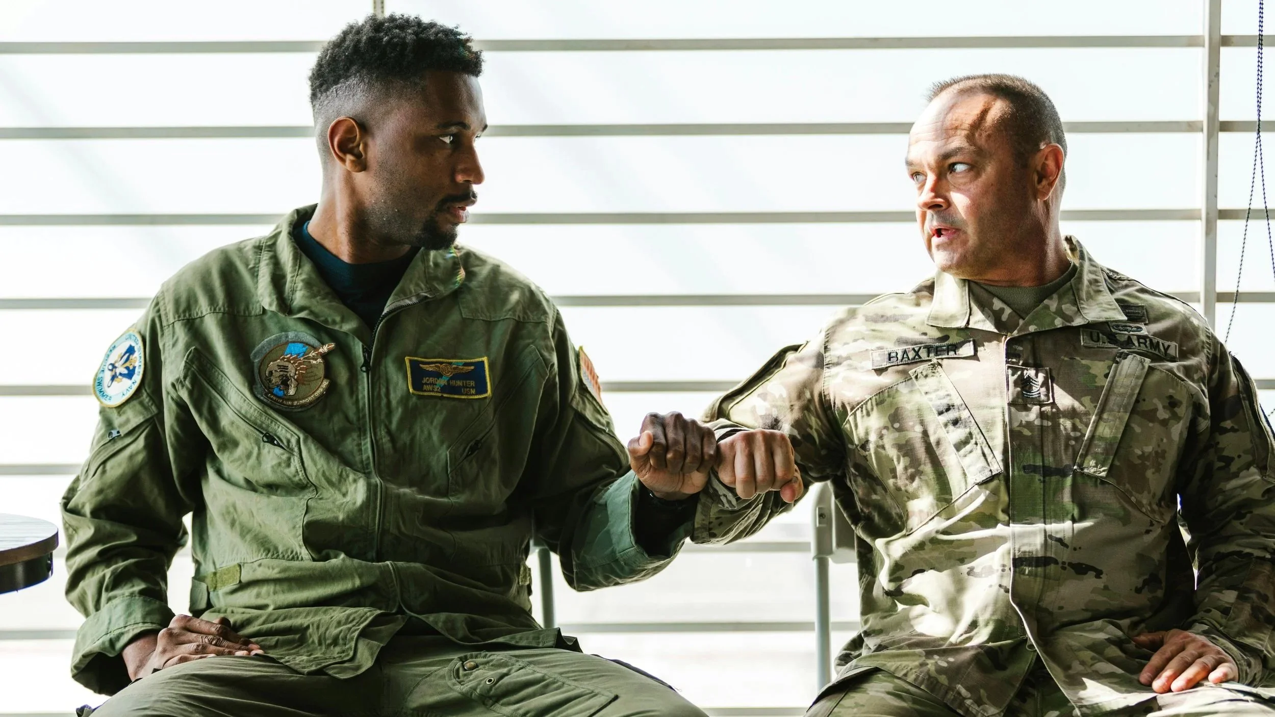 Two men sitting in military fatigues fistbumping; the man on the left is Black and the man on the right is white