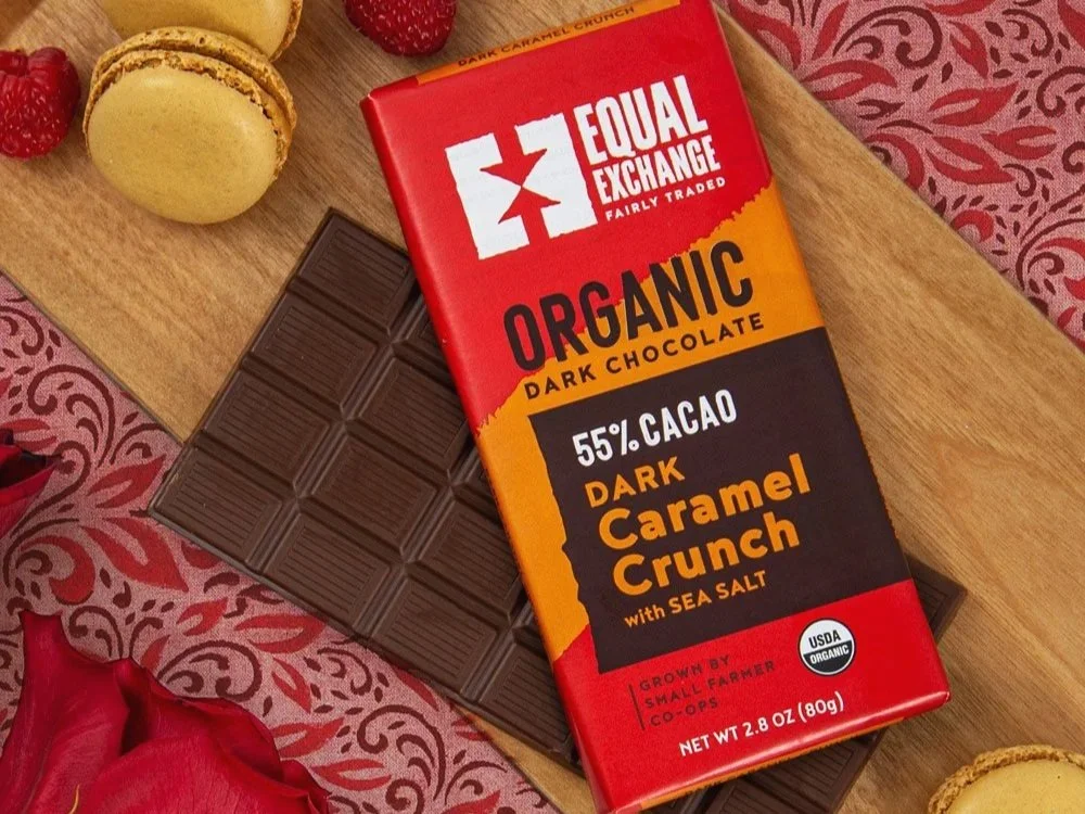 Chocolate bar from Equal Exchang with a red wrapper sits on a cutting board, surrounding by macarons, raspberries, and roses