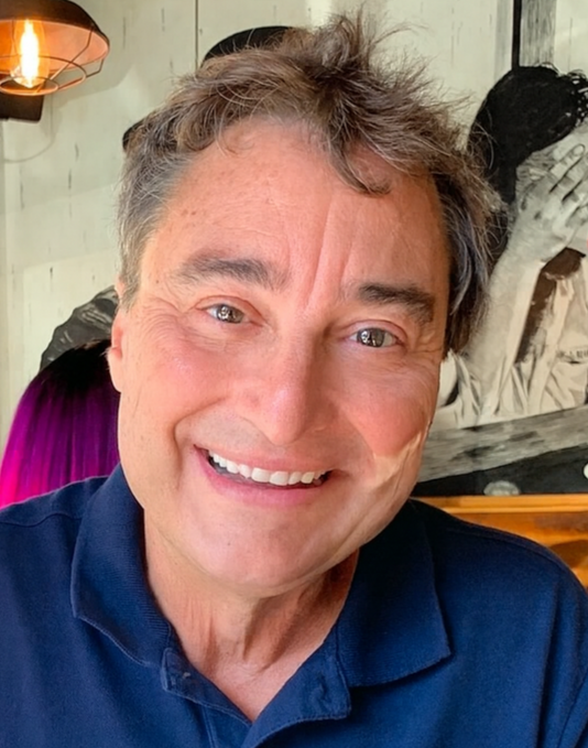 Smiling man with gray hair wearing a dark blue shirt in a cozy, decorated room.