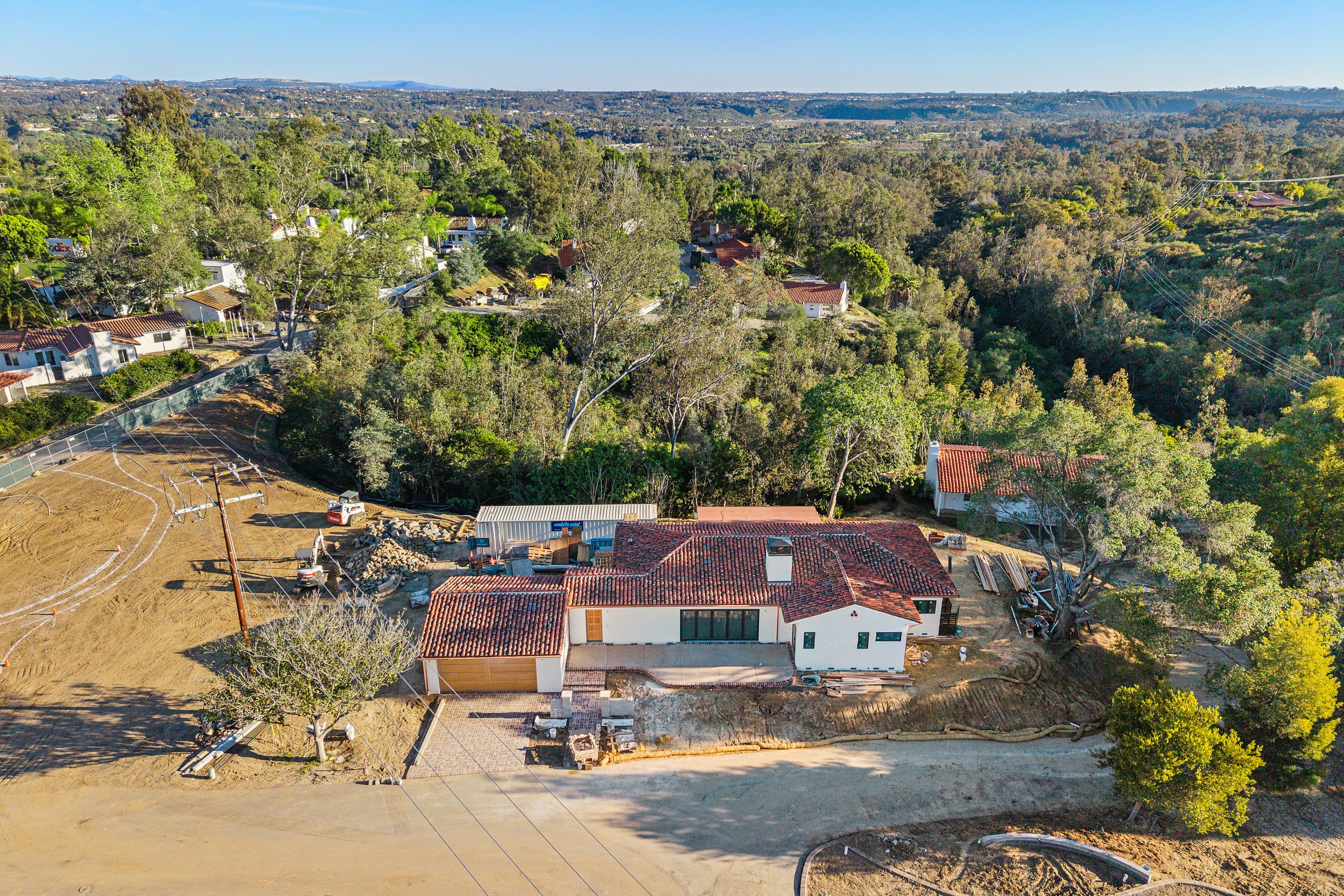 Aerial view of a residential area with a mix of finished houses and houses under construction, surrounded by lush green trees and hills in the background.